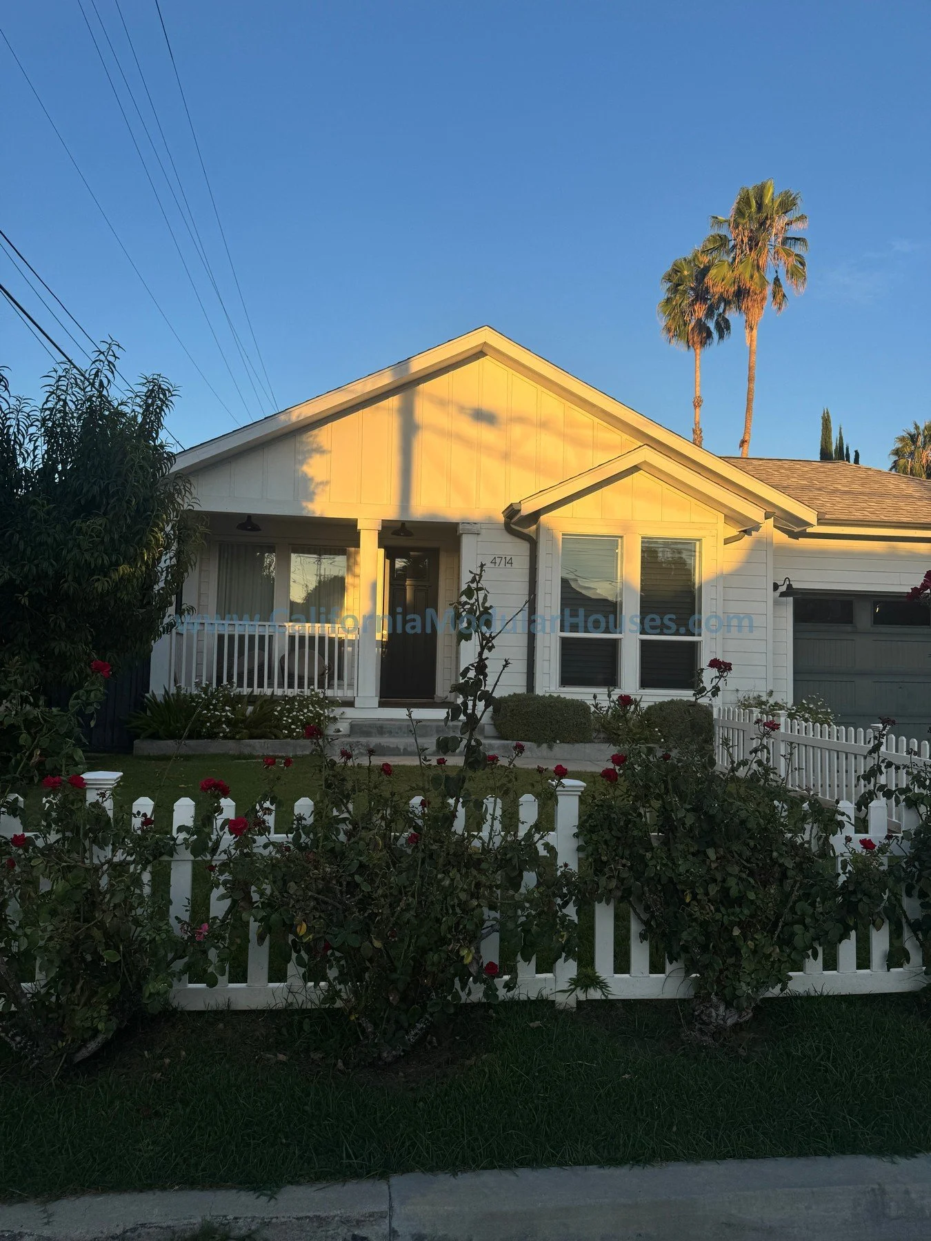 Front view of a white house with a small front porch, surrounded by a white picket fence and a garden with rose bushes, under a clear blue sky with palm trees in the background.