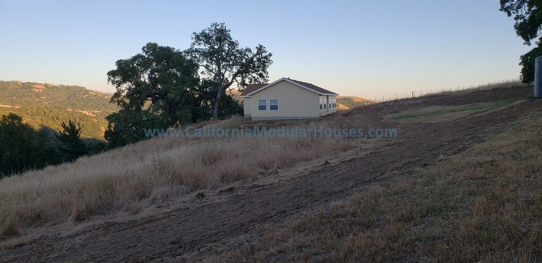 A small yellow house on a hill with a dirt path beside it, surrounded by trees and hills in the background, with a water tank on the far right.  San Jose Modular Home, Modular Homes California,