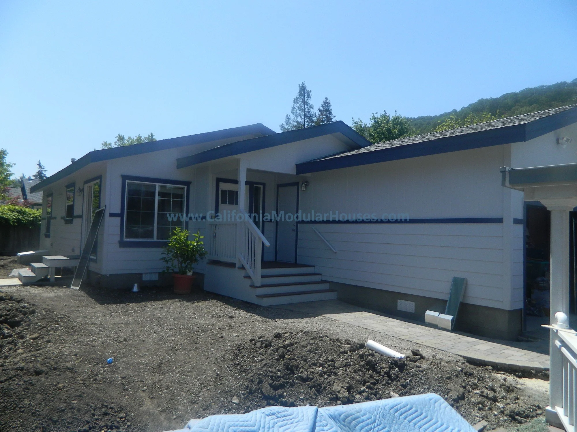 A white house under construction with steps leading to a small porch, surrounded by dirt and construction materials, with a backdrop of trees and blue sky.
