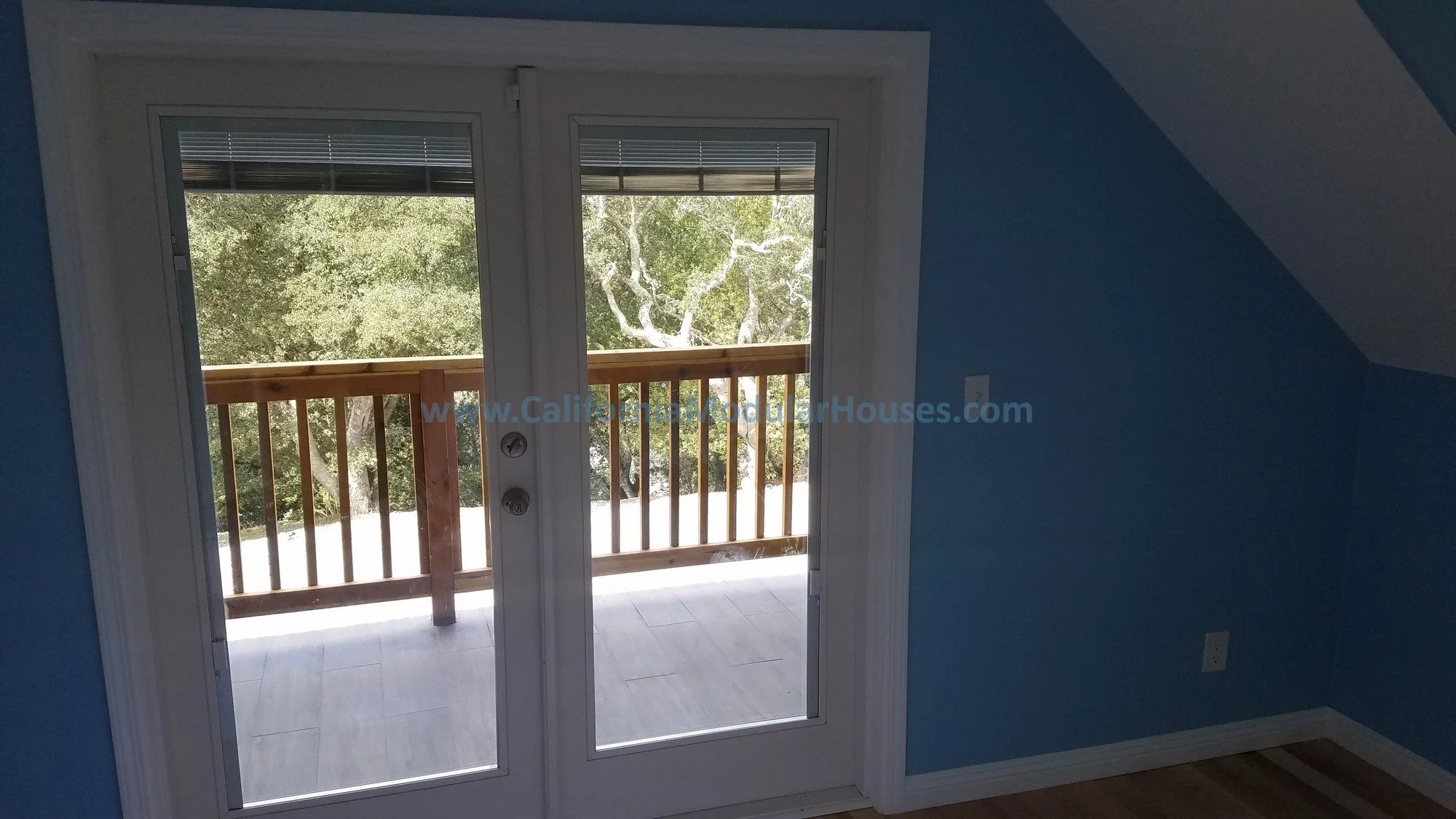 Interior view of a room with blue walls, a door with glass panels leading to a balcony with a wooden railing, and trees visible outside.  This is from the attic showing the site completed attic, and the French Doors with the covered porch. 