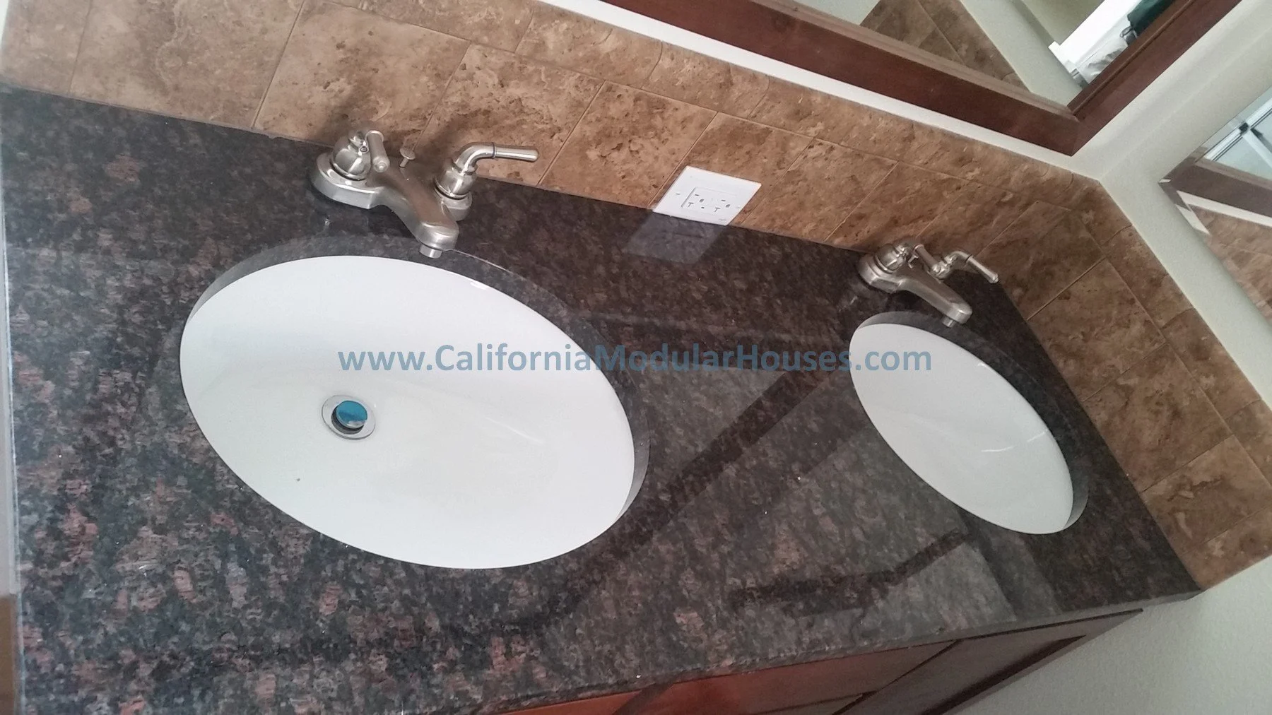 Double bathroom vanity with two white oval sinks and silver faucets on a dark granite countertop, with a brown-tiled wall behind and a mirror reflecting part of the room.  Interior image of Accessory Dwelling Unit Morgan Hill, Santa Clara County, CA