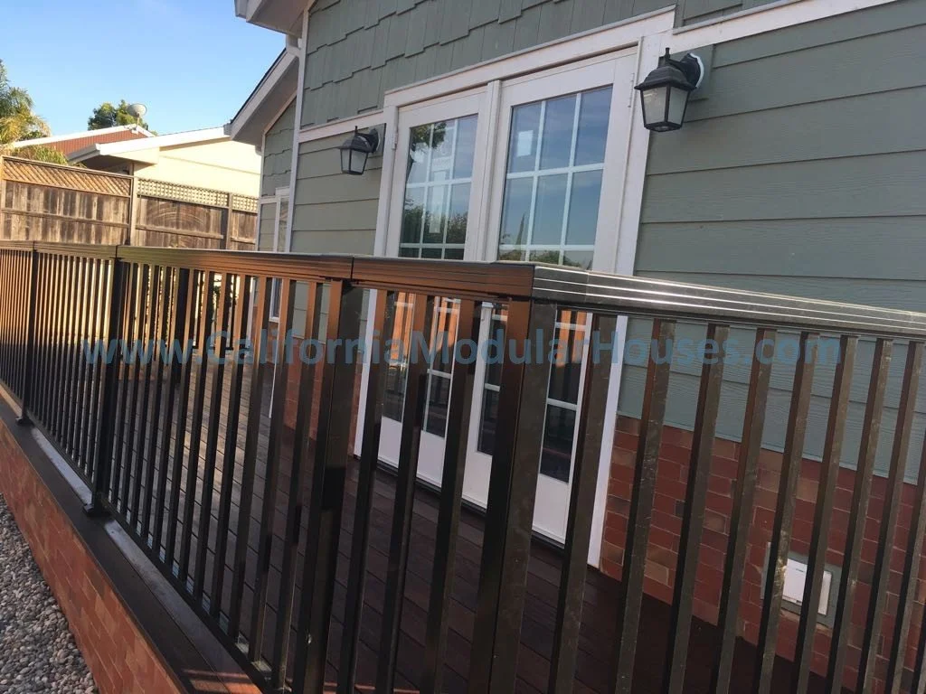 A residential house balcony with black metal railing, green siding, brick foundation, white-framed window and door, and two outdoor wall-mounted lamps.