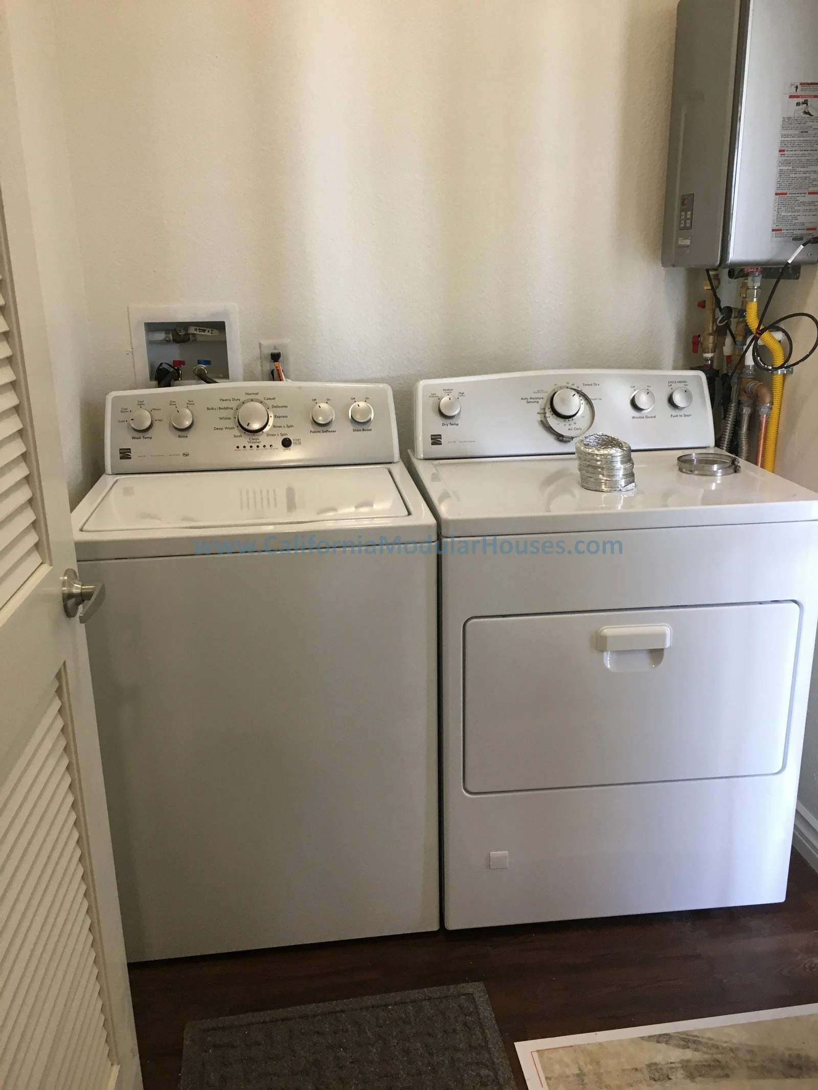 A white washing machine and dryer side by side in a laundry room, with a small window or control panel above each appliance. The dryer has a duct or vent pipe at the top with a shiny metallic cover.