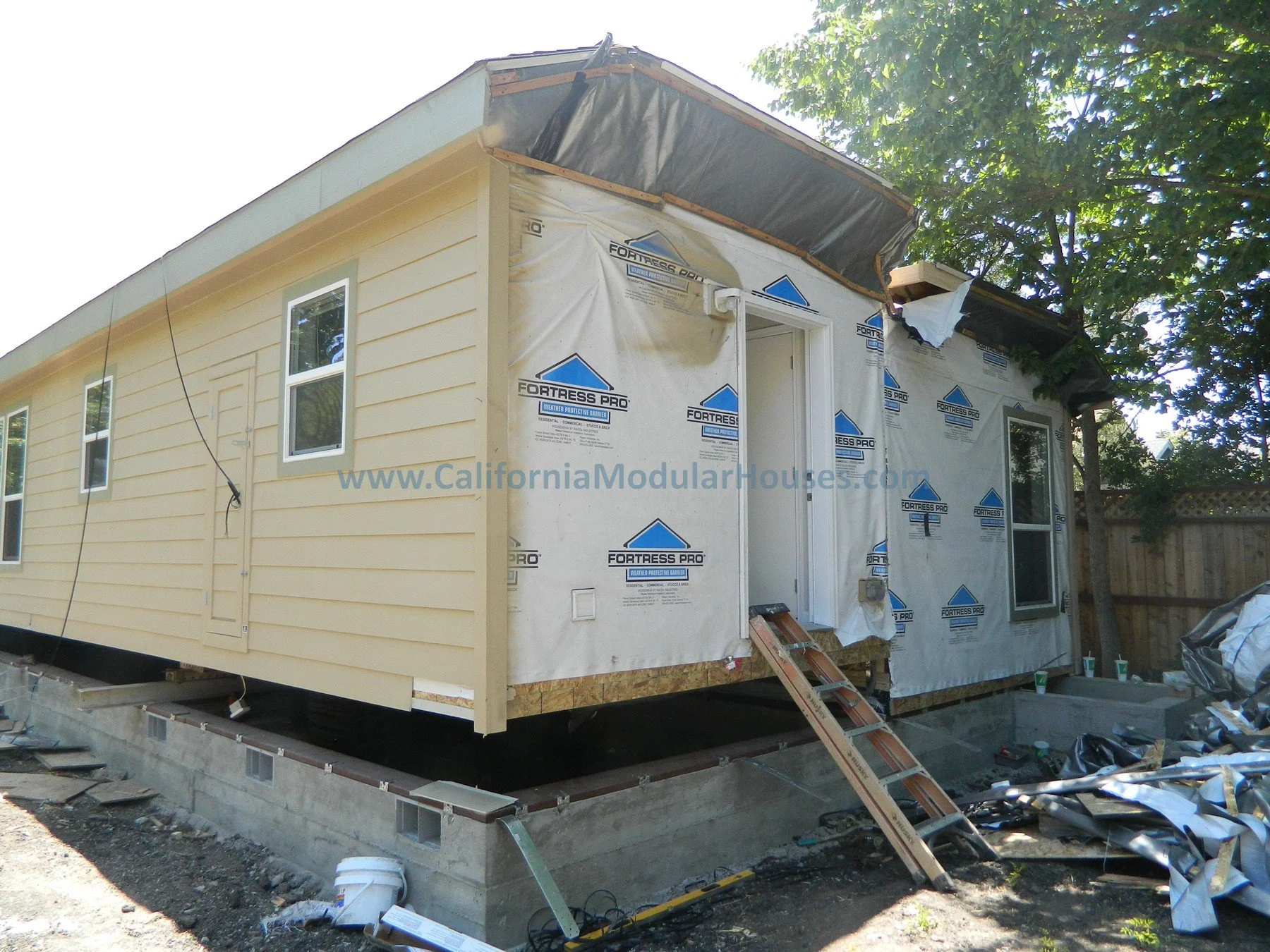 A house under construction showing siding being installed, with windows installed, and a construction ladder leaning against the house. The house is on a concrete foundation, and building materials are visible nearby.