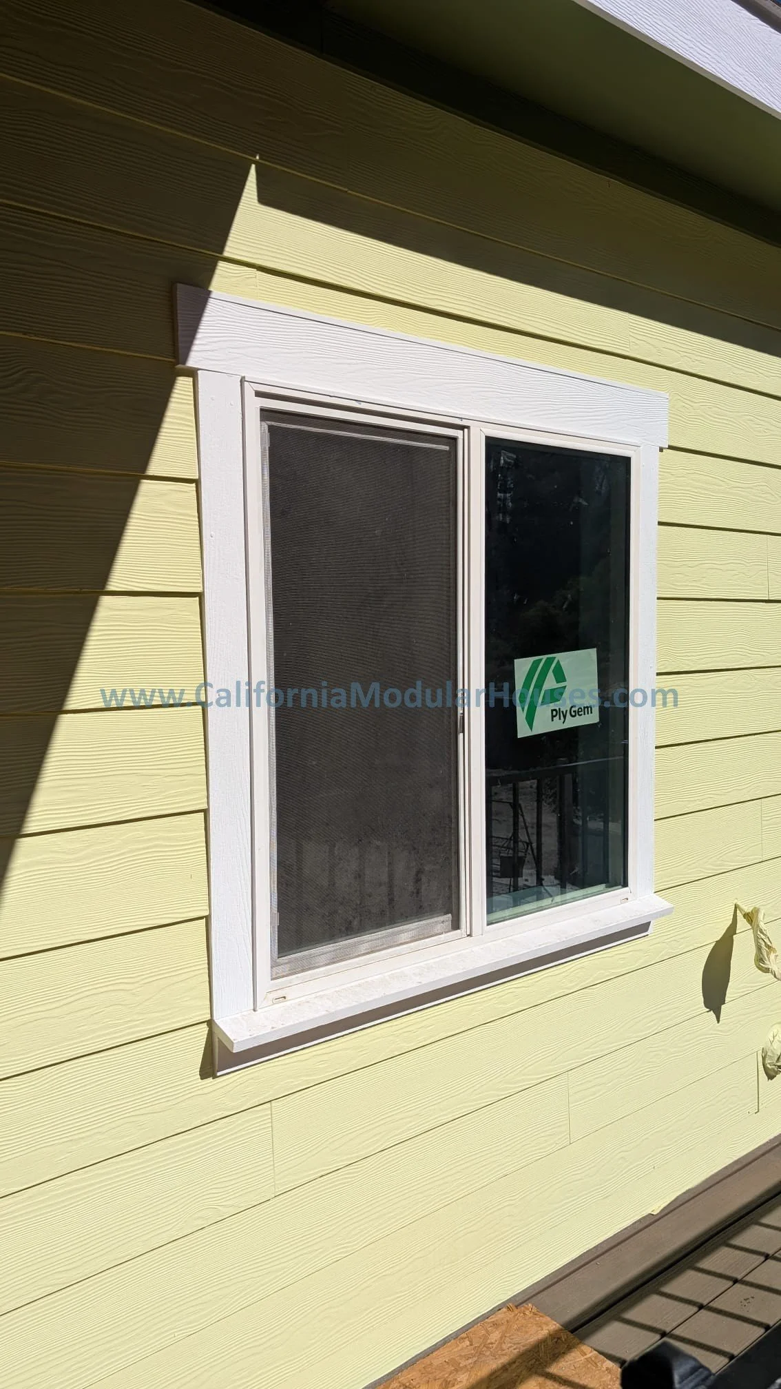 Photo of a window with white trim on a yellow siding house. The window has a screen and a sticker that reads 'Ply Gem'.