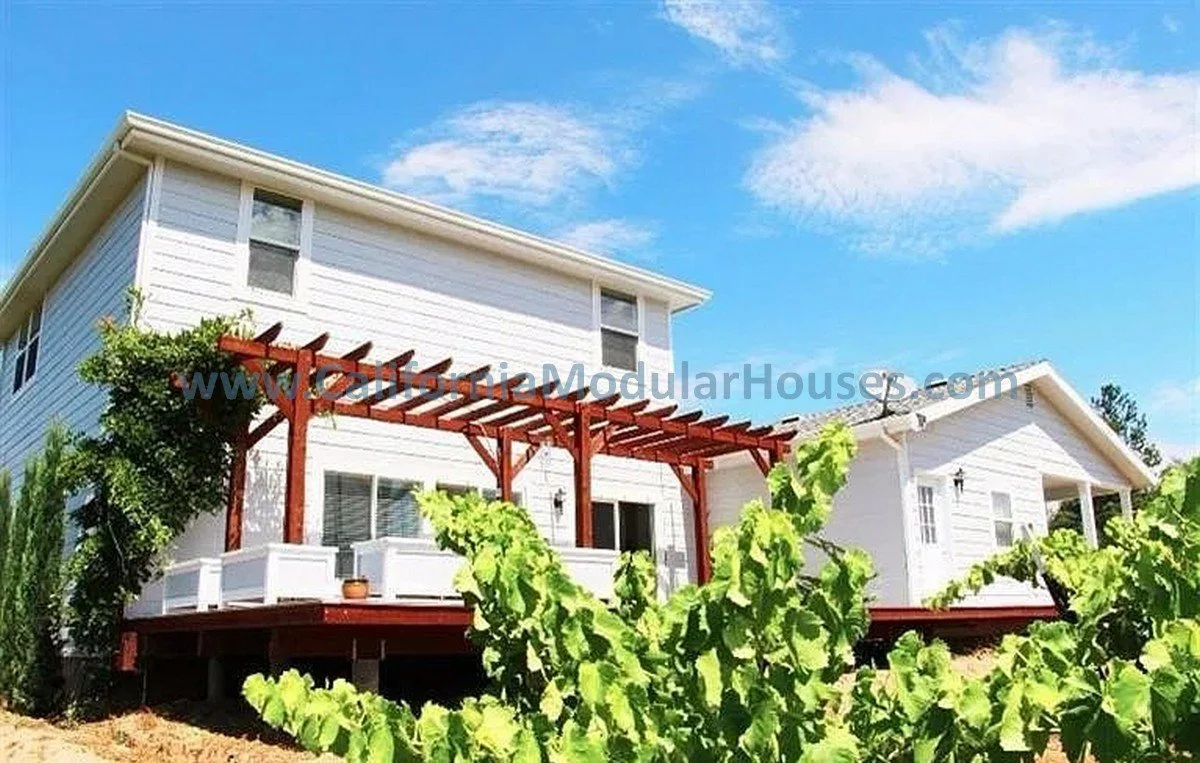 White two-story modular house with a wooden pergola on the back porch, surrounded by green plants and a bright blue sky with a few clouds.