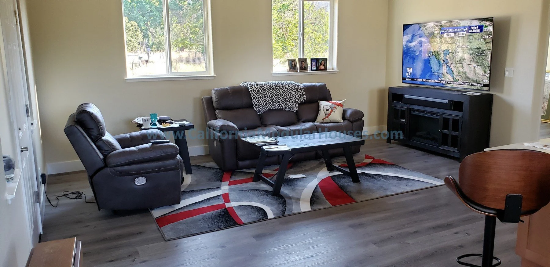 Living room with a black reclining chair and a sofa, a rectangular coffee table with magazines, a TV on a black stand, a colorful rug, two windows with trees outside, and a small side table with cups and papers.