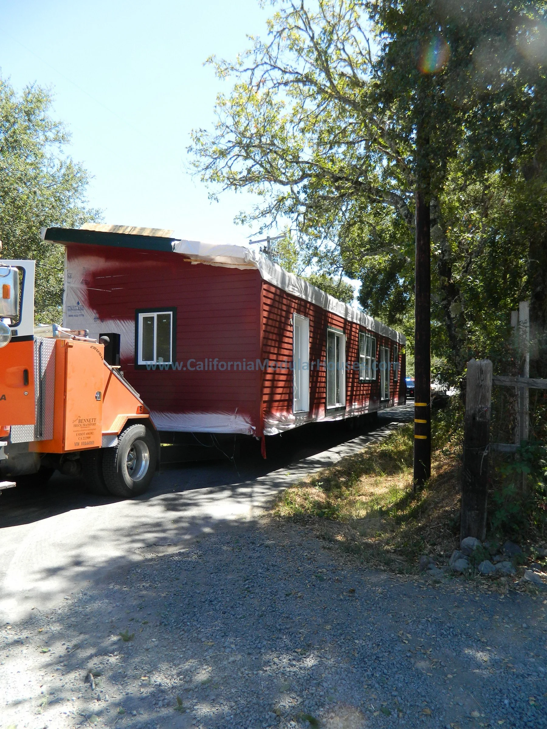 A red modular house being transported on a flatbed truck on a gravel road, with trees and a utility pole nearby under a clear blue sky.   California Modular Houses, Middletown Prefab Homes.
