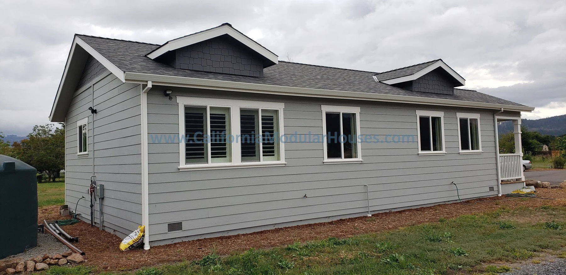 Side view of a newly built gray house with white trim, multiple windows with shutters, a small front porch, and a gabled roof, situated in a semi-rural area with cloudy skies.    2 bedroom, 2 bathroom modular ADU from the exterior.   