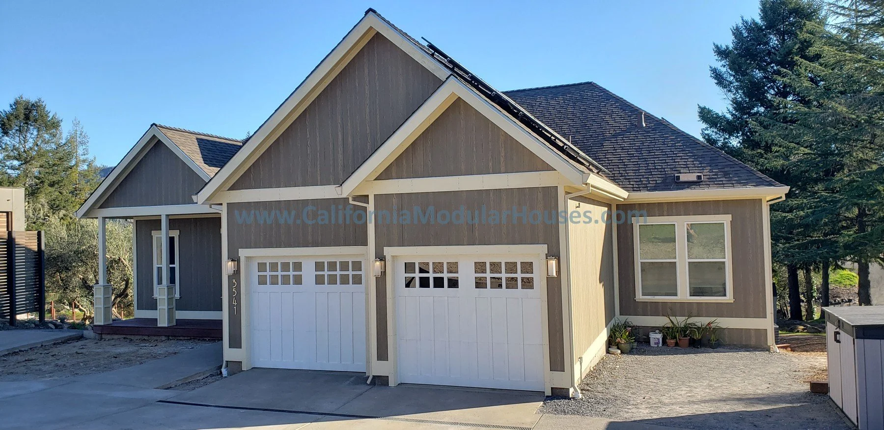A new modular house with gray exterior walls, white trim, and two white garage doors, situated in a sunny outdoor setting with trees in the background.  Santa Rosa, CA.  Sonoma County.  Modular Homes Northern California, Prefab Houses, Prefab Home CA