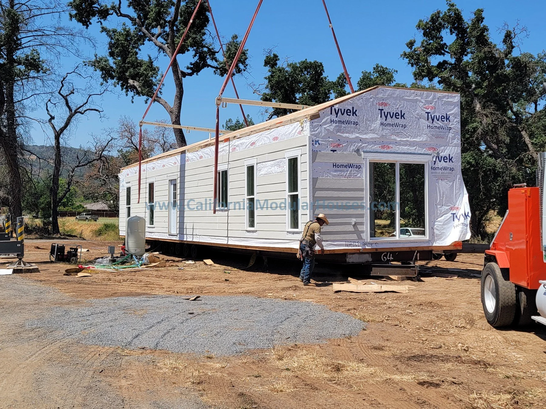 A worker stands next to a prefabricated house under construction, with partially finished exterior walls, window openings, and construction equipment around, situated on a dirt lot with trees in the background.