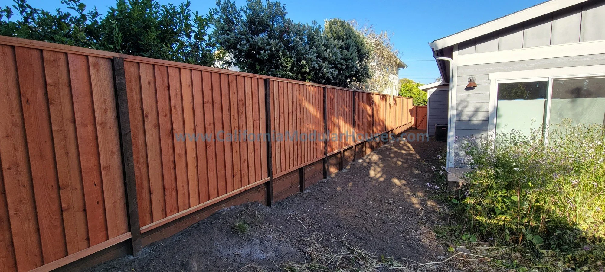 Backyard with newly installed wood privacy fence along the yard's perimeter, next to a house with an outdoor wall-mounted light, and a garden bed with plants and flowers, under a clear blue sky.