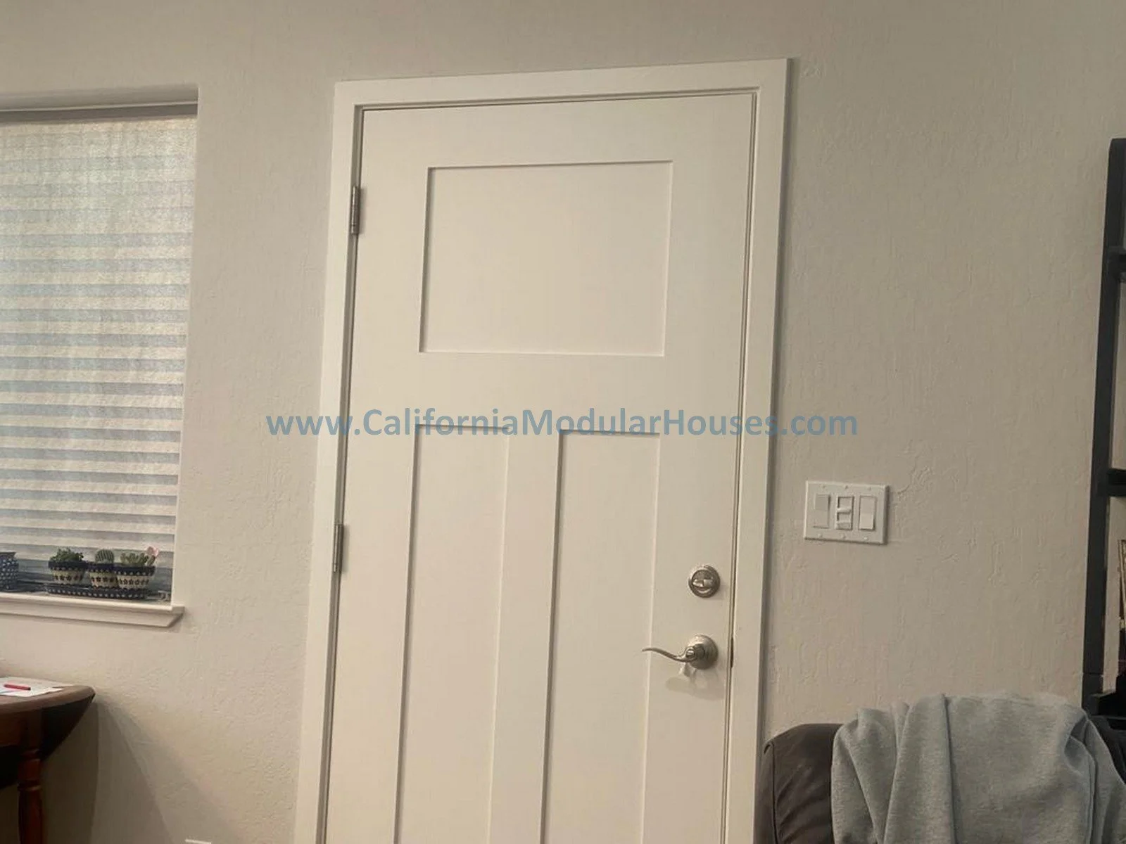 Interior view of a white door with a door knob and deadbolt lock, next to a window with closed blinds, a light switch on the wall, and part of a table with a small plant on it.