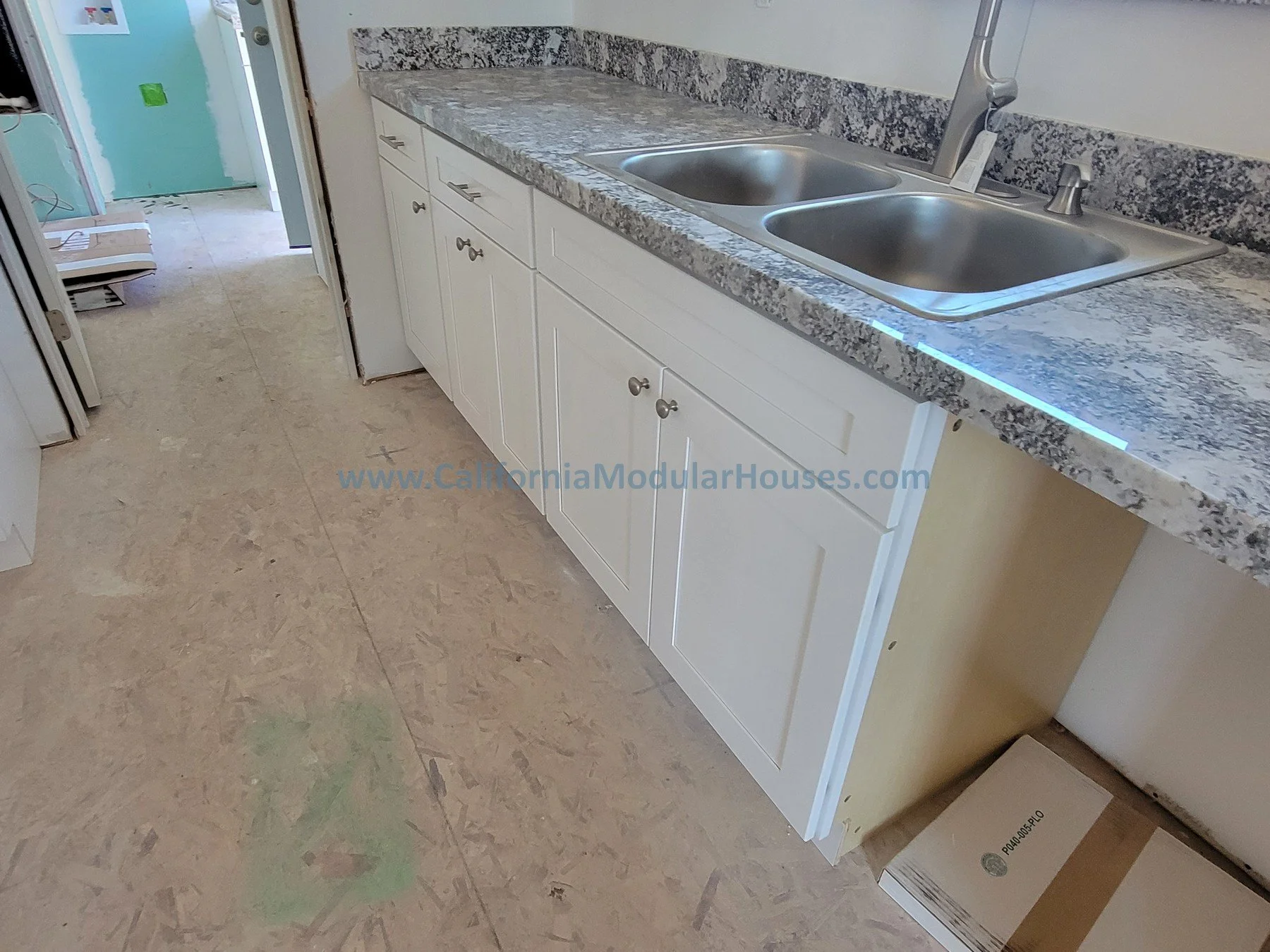 Kitchen with granite countertop, double stainless steel sink, white cabinet doors, and unfinished flooring.