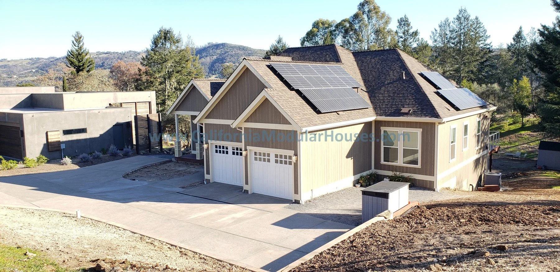 A modern two-story house with grey siding and solar panels on the roof. The house has two white garage doors and is surrounded by a concrete driveway and landscaped yard. There are trees and hills in the background. Modular Homes, Prefab Homes.