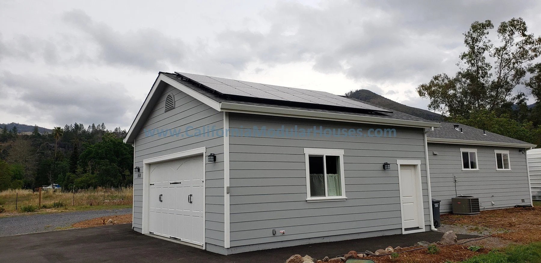 A light gray modular house with a dark shingled roof equipped with solar panels, a white garage door, a side door, two small windows, and outdoor wall-mounted lights.  Kenwood, Sonoma County, CA. Backyard home.  