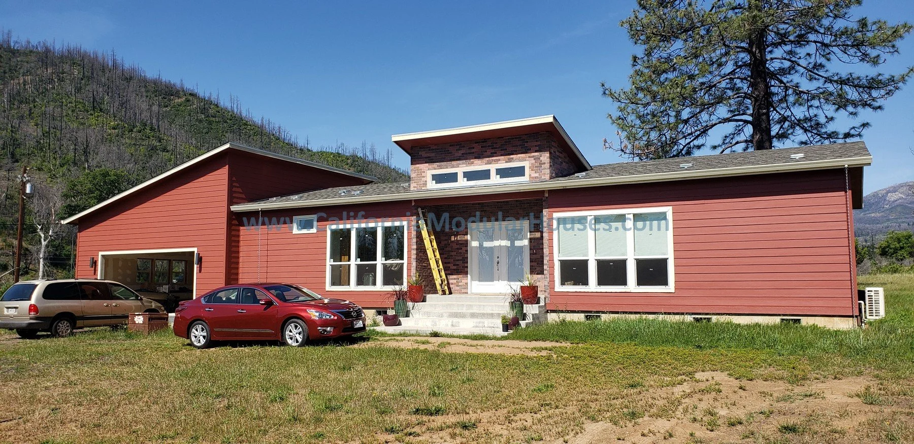 Newly constructed modern house with pink siding, large windows, and a brick entryway, surrounded by a grassy yard, set against a mountainous background and blue sky. Lake County Modular, Northern CA Modular Home.