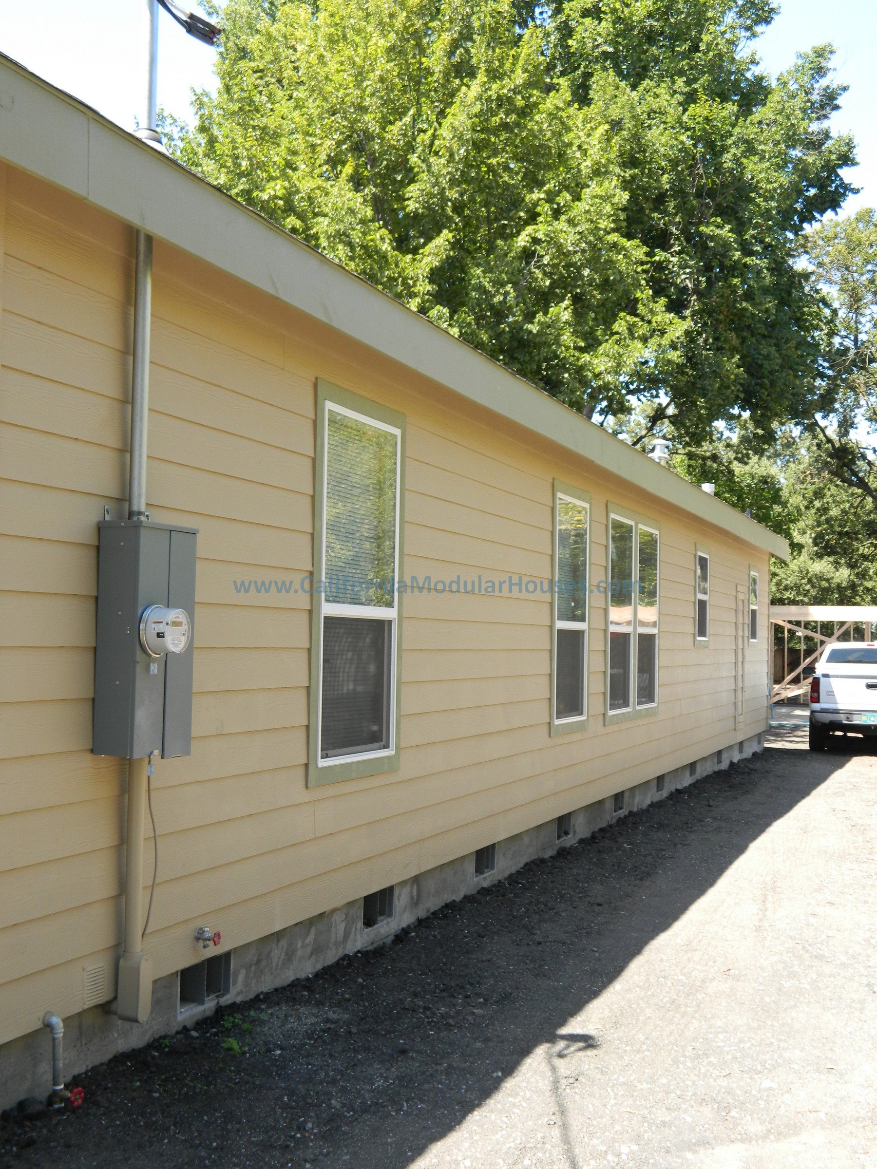 Exterior view of a beige modular home with four vertical windows, electrical meter, and conduit pipes, situated on a gravel ground with a white truck parked nearby and green trees in the background.