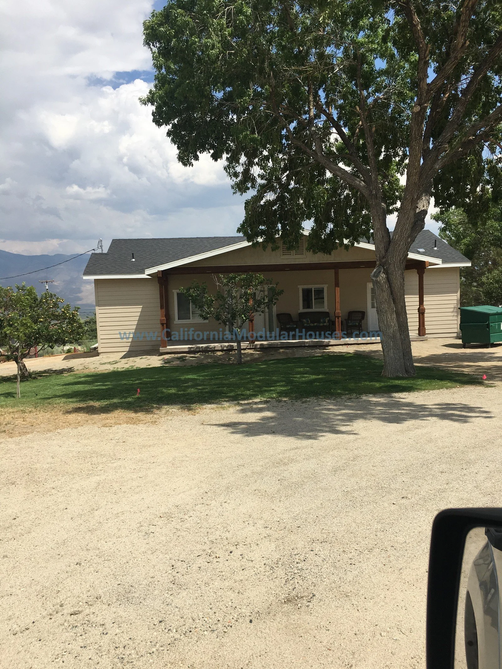 A beige modular house with a small front porch, surrounded by trees and green grass, with a dirt driveway and mountains in the background.