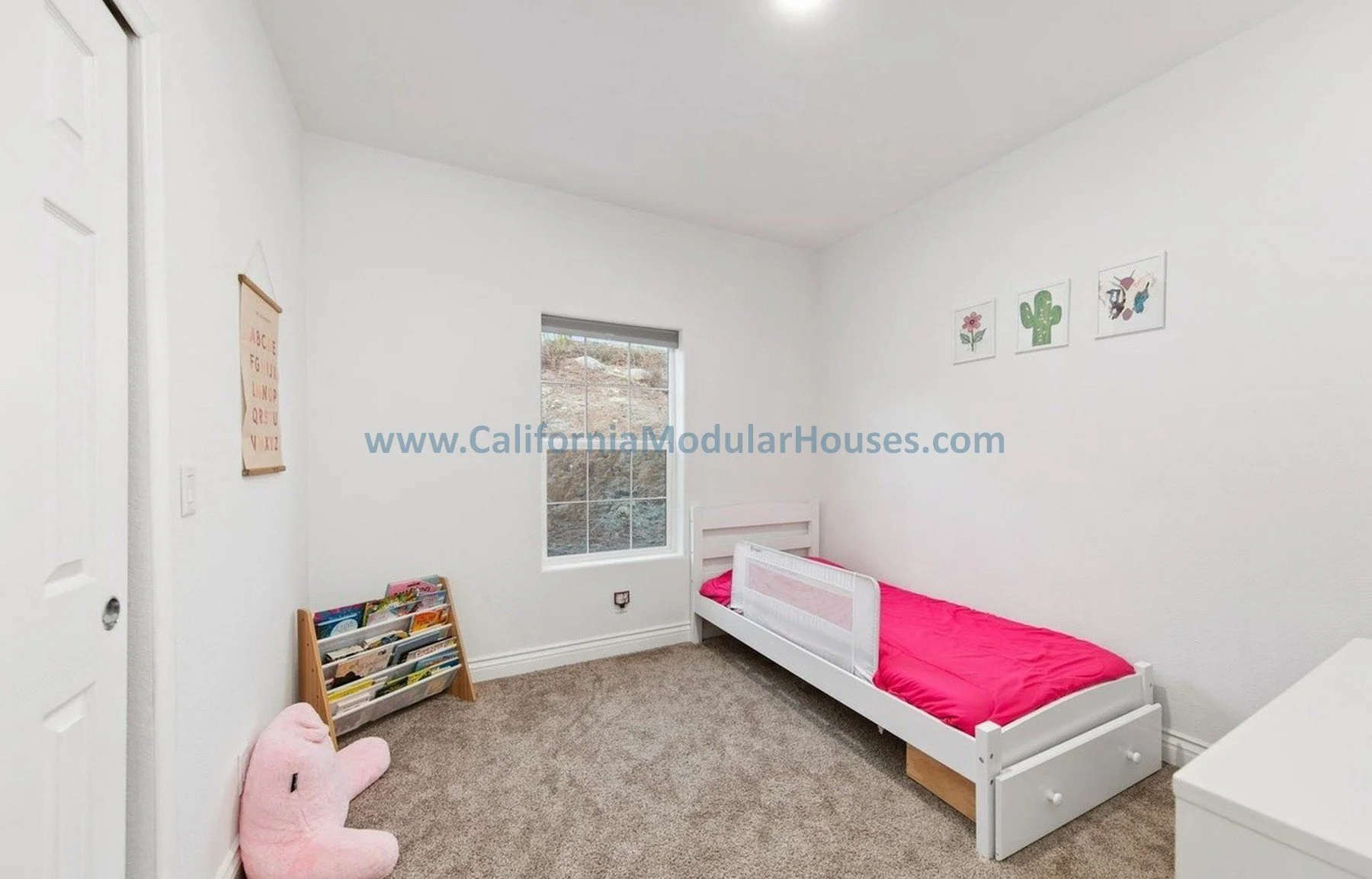 A small children's bedroom with white walls and ceiling, beige carpet, and a single window with a view of dirt and rocks outside. It has a white bed with a pink mattress and pillow, located along one wall. On the wall above the bed are three framed p