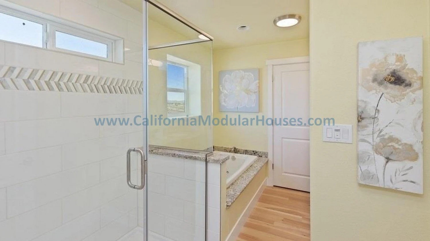 Bathroom with a glass shower, bathtub, window, beige walls, wooden floor, and floral artwork.  Modular house interior.   PreFab Modular for California.