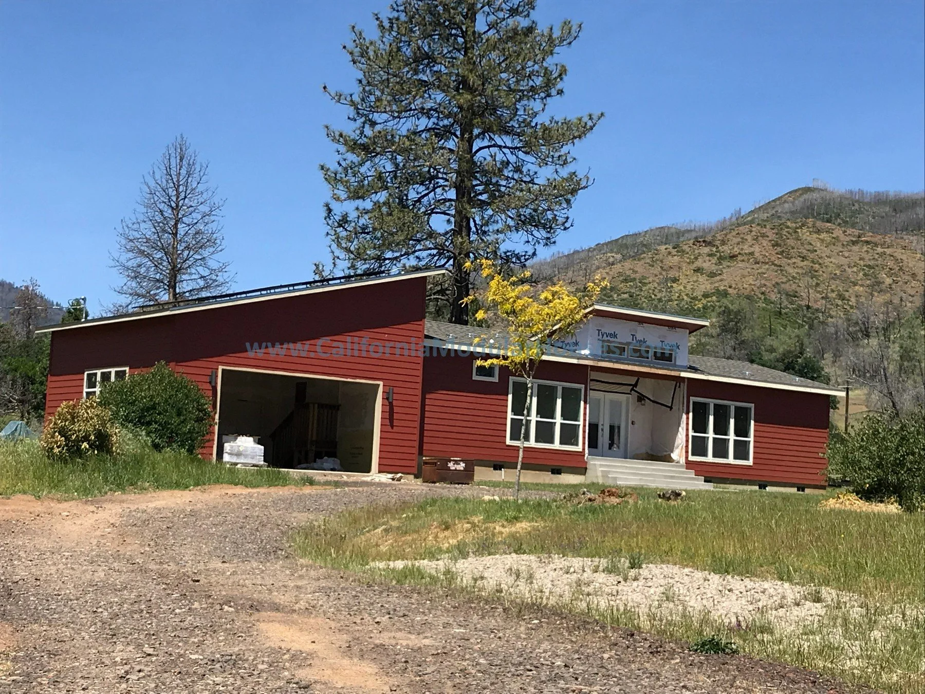 Modular Homes, Modular Homes California. A house under construction with red exterior siding, a garage, and a front entrance with steps. The house is surrounded by grass and trees, with a mountain in the background. 