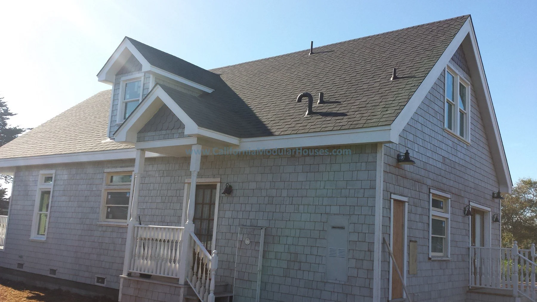 A two-story house with gray shingle siding, a gable roof, and multiple windows. The front porch has a small set of stairs with white railings. The house has a dormer window on the roof. Mendocino City, CA. Mendocino County, California. Prefab modular