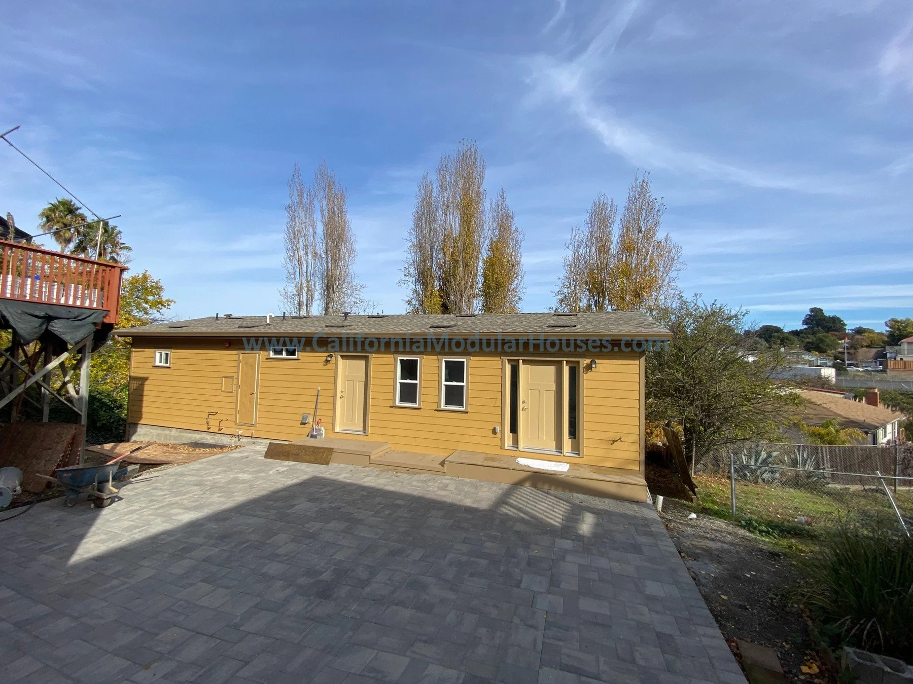 Newly constructed small yellow modular house with a flat roof, three windows, and two doors, situated on a paved outdoor area. Construction materials and tools are present nearby, and the sky is clear with a few clouds, with trees in the background.