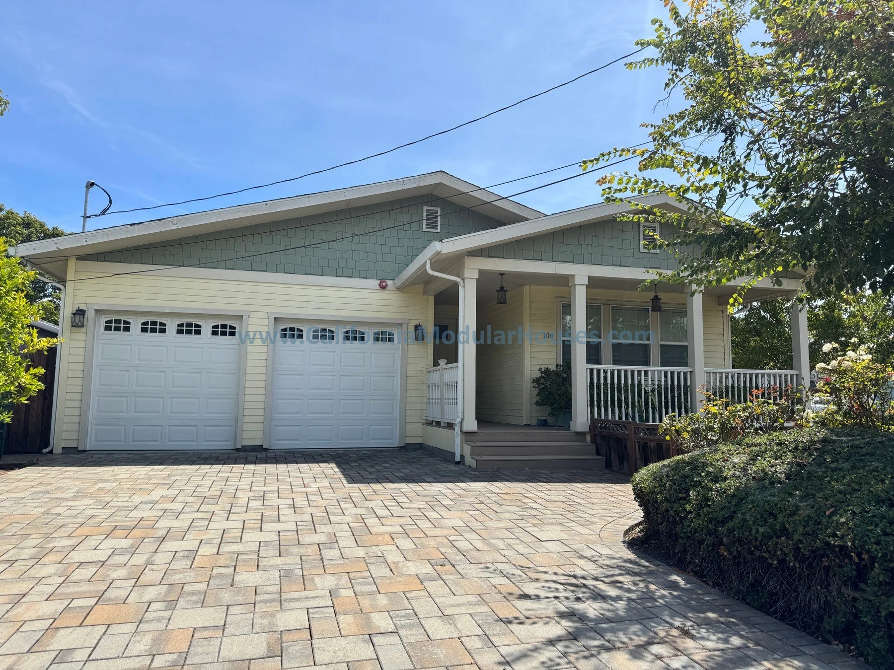 Front view of a two-story house with yellow and blue-gray siding, a white garage door, a porch with white railings, and a landscaped yard with bushes and trees.