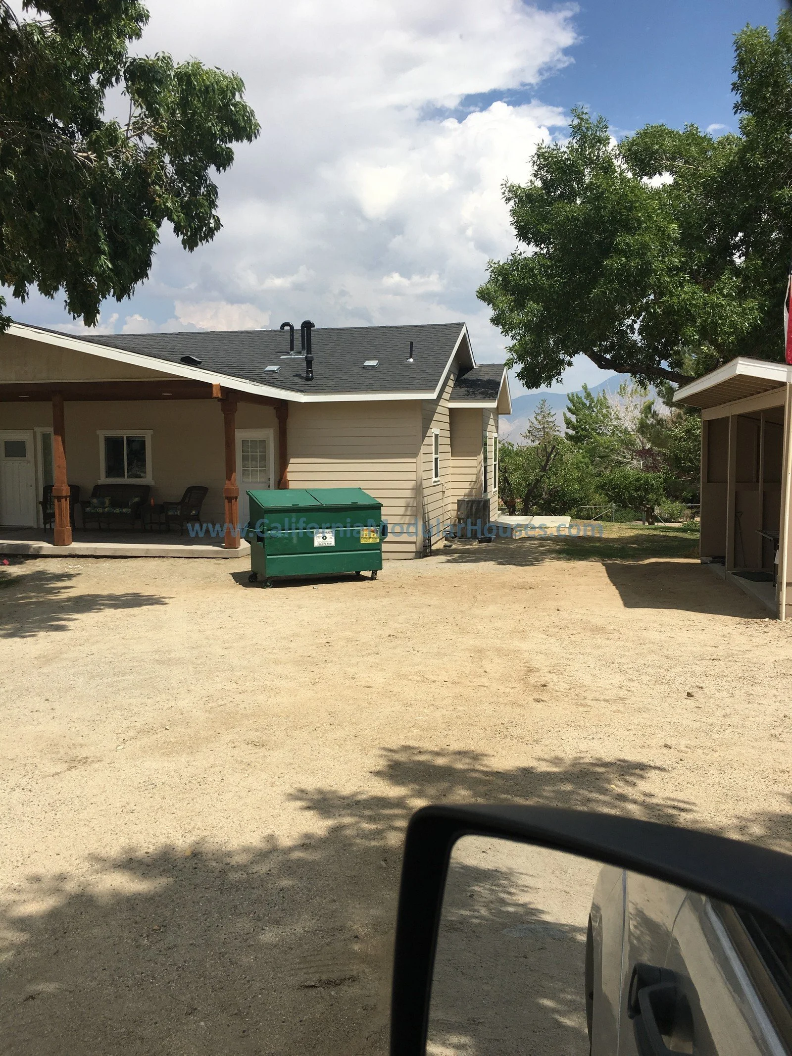 Residential backyard with a beige house, a green trash bin, a covered patio with outdoor furniture, large trees, and a dirt ground under a partly cloudy sky.