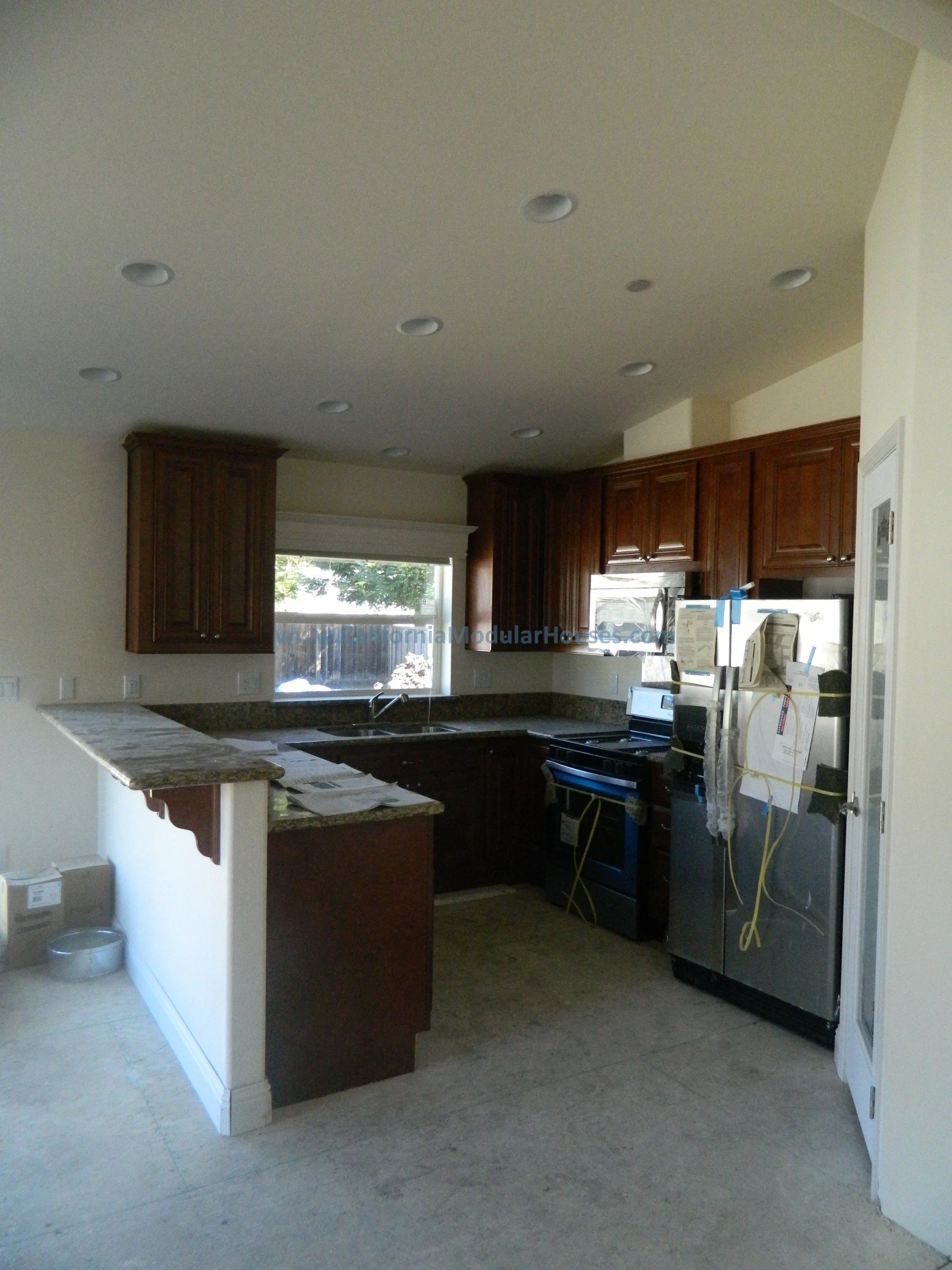 Unfinished kitchen with dark wood cabinets, granite countertops, stainless steel appliances, and a window above the sink, with some papers and wires on the refrigerator.