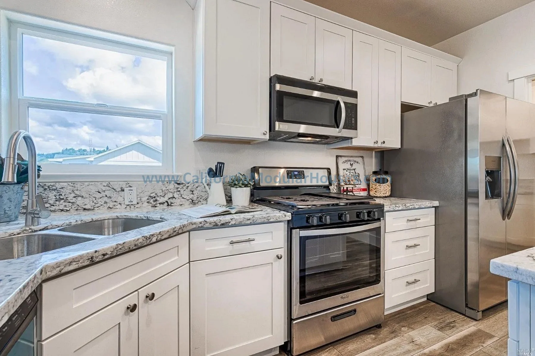 Modern kitchen with white cabinets, granite countertops, stainless steel appliances including refrigerator, oven, microwave, and dishwasher, under a window with a view of a cloudy sky and neighboring rooftops.