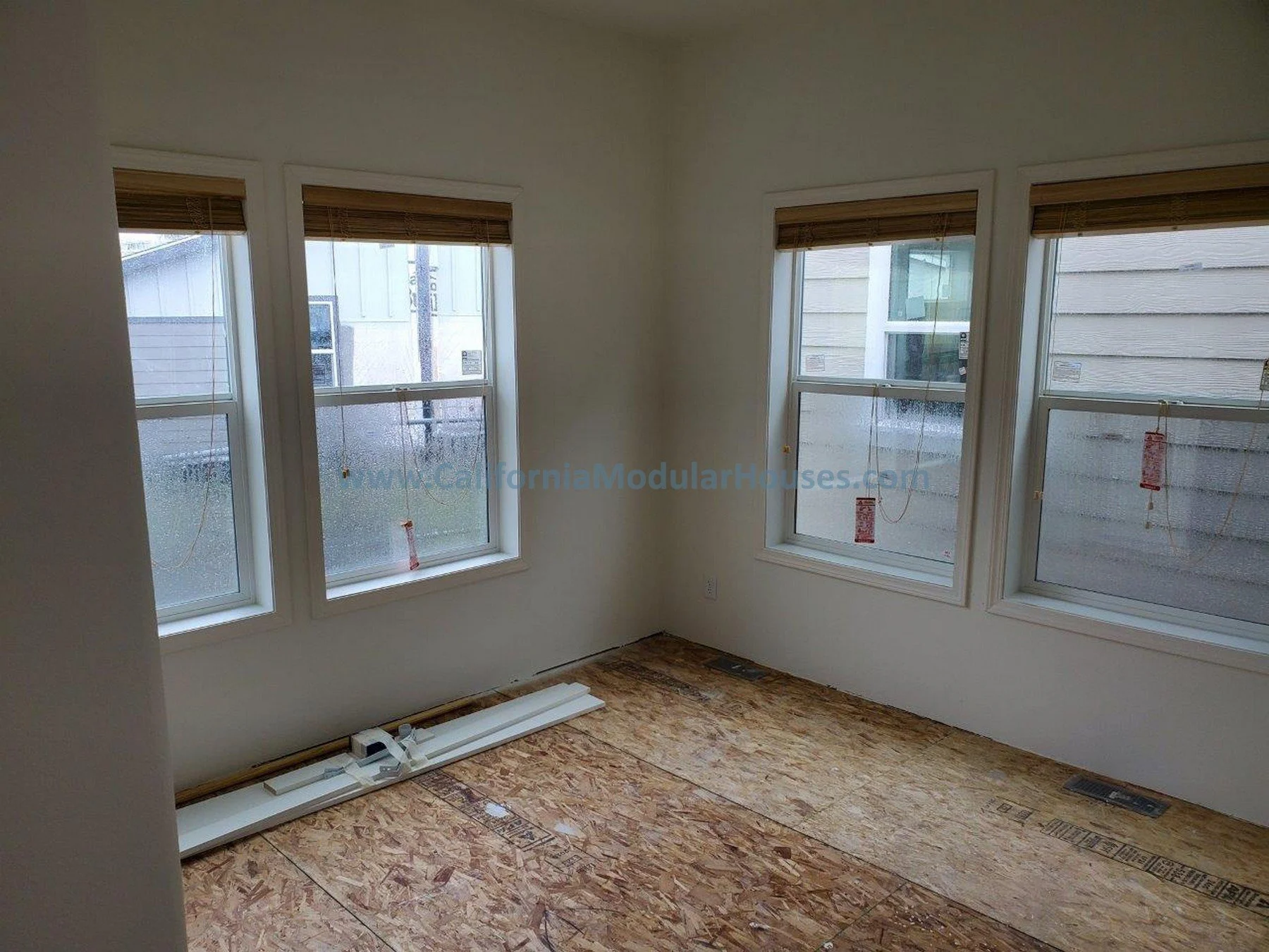 Empty room with four windows showing rain outside, wooden blinds, and partially installed baseboard heating on a plywood floor. Modular Home CA, California Modular Homes, Prefab Modular Homes California.
