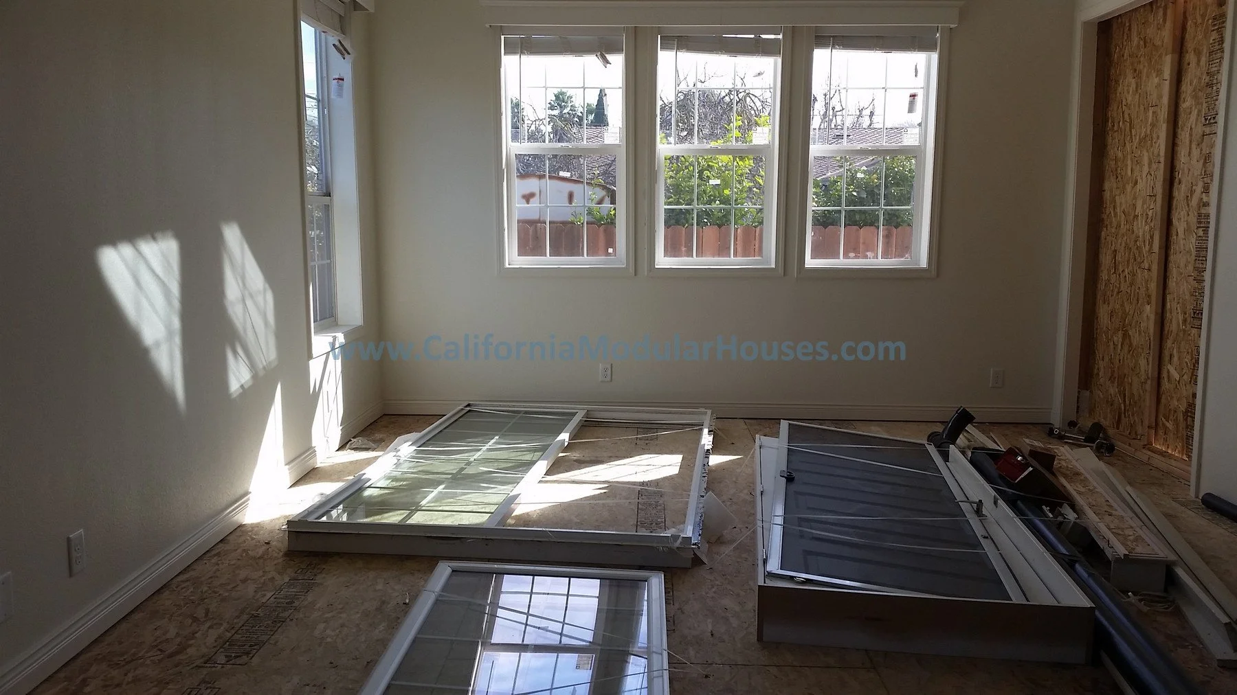 Room under renovation with disassembled window frames on the floor and three large windows on the wall showing trees and a fence outside. Bay Area Prefab Modular Home, California Modular Homes,