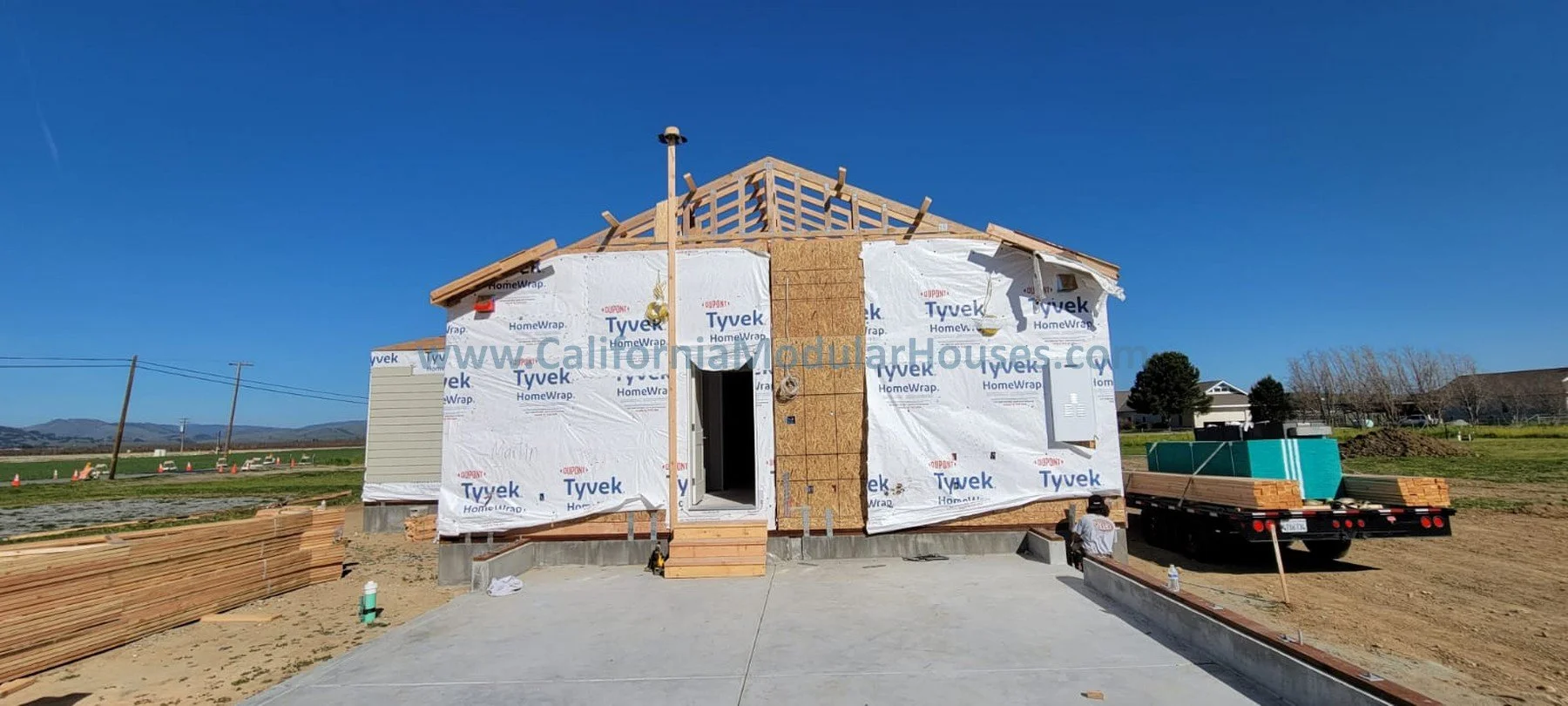 A house under construction with wooden framing and exterior insulation, on a concrete foundation with a new driveway, in a rural area with open fields and a blue sky.