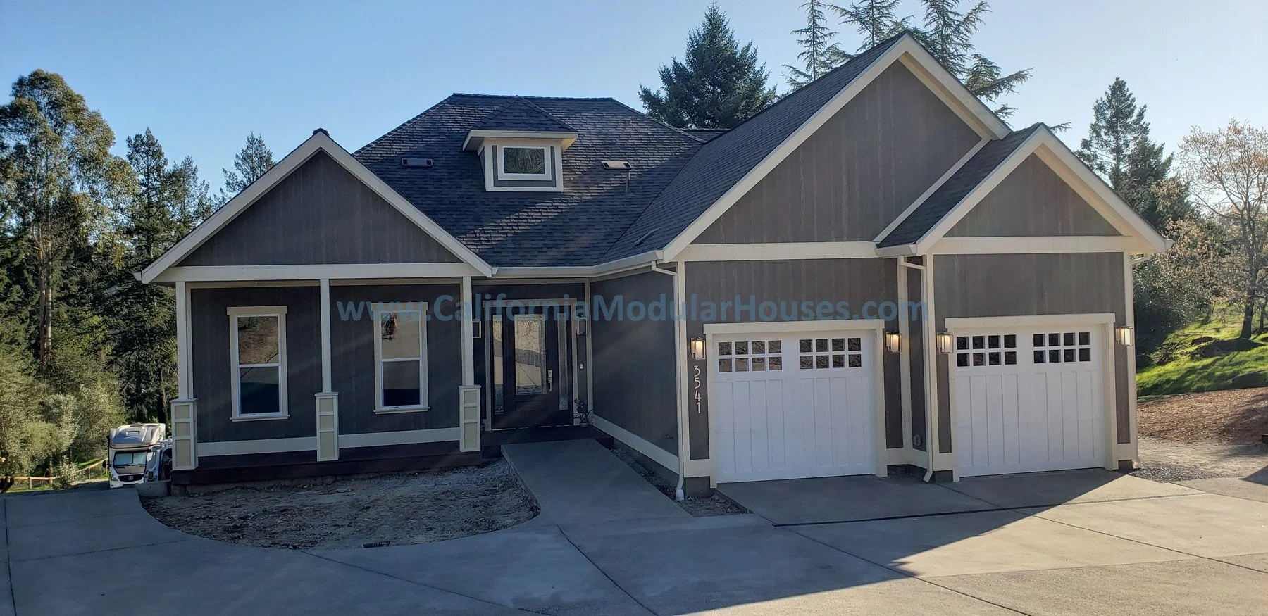 Front view of a modern gray modular house with a two-car garage, white trim, and a front porch, surrounded by trees and a concrete driveway.  Modular Home CA, Modular Homes Bay Area, Pre-Fabricated Homes, Santa Rosa, CA.  Sonoma County.  