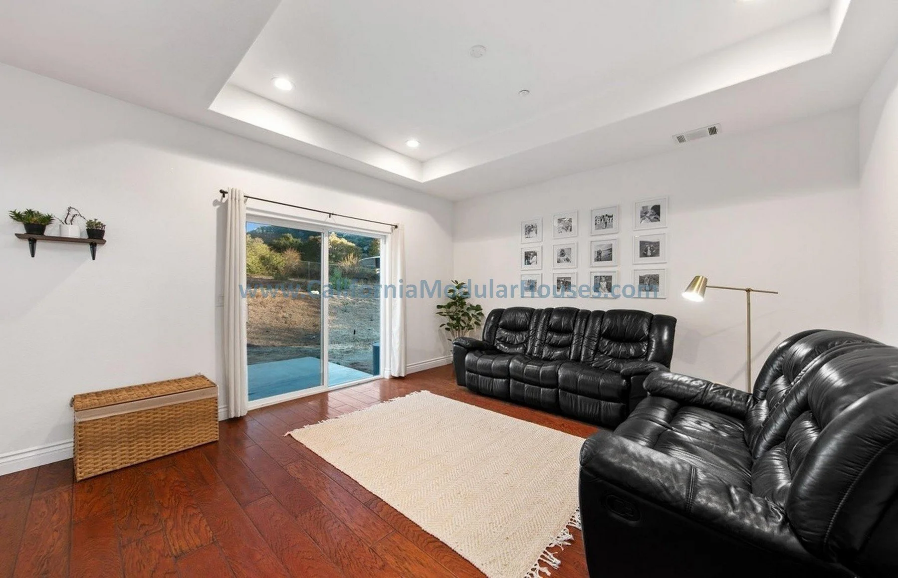 Living room with white walls, hardwood floors, black leather sofas, a small woven basket, a window with white curtains, wall art, and a modern floor lamp.