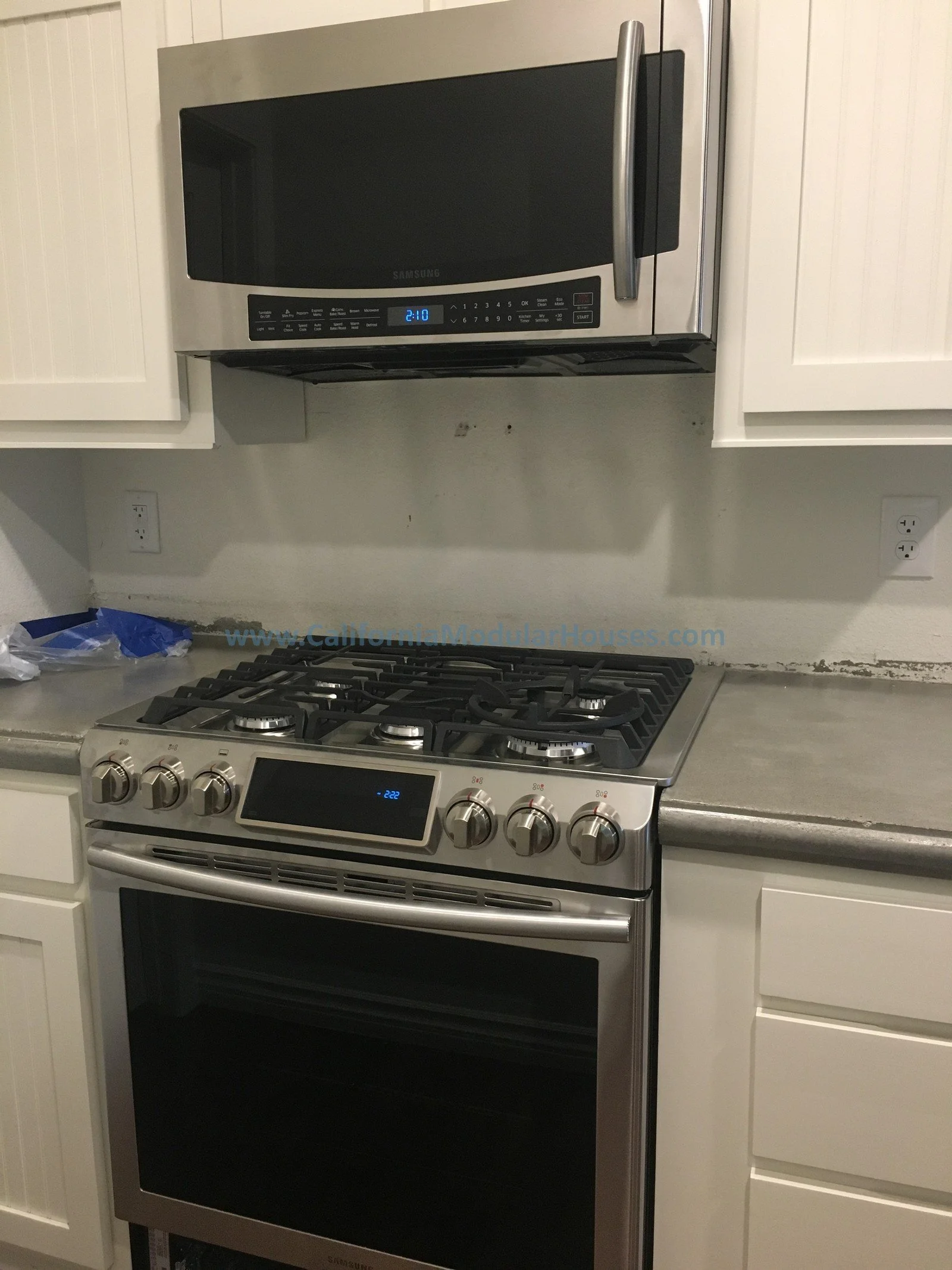 Stainless steel microwave oven above a stainless steel gas stove with an oven, in a kitchen with off-white cabinets and a gray countertop.