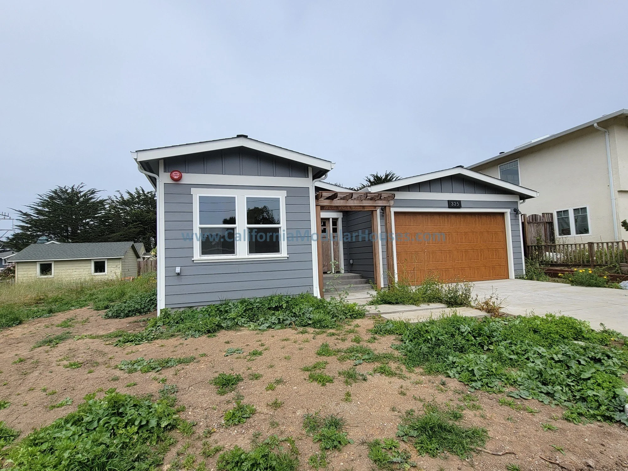 A two-story house with gray siding and a wooden garage door, with a small garden and dirt yard in the front, and neighboring houses visible in the background.
