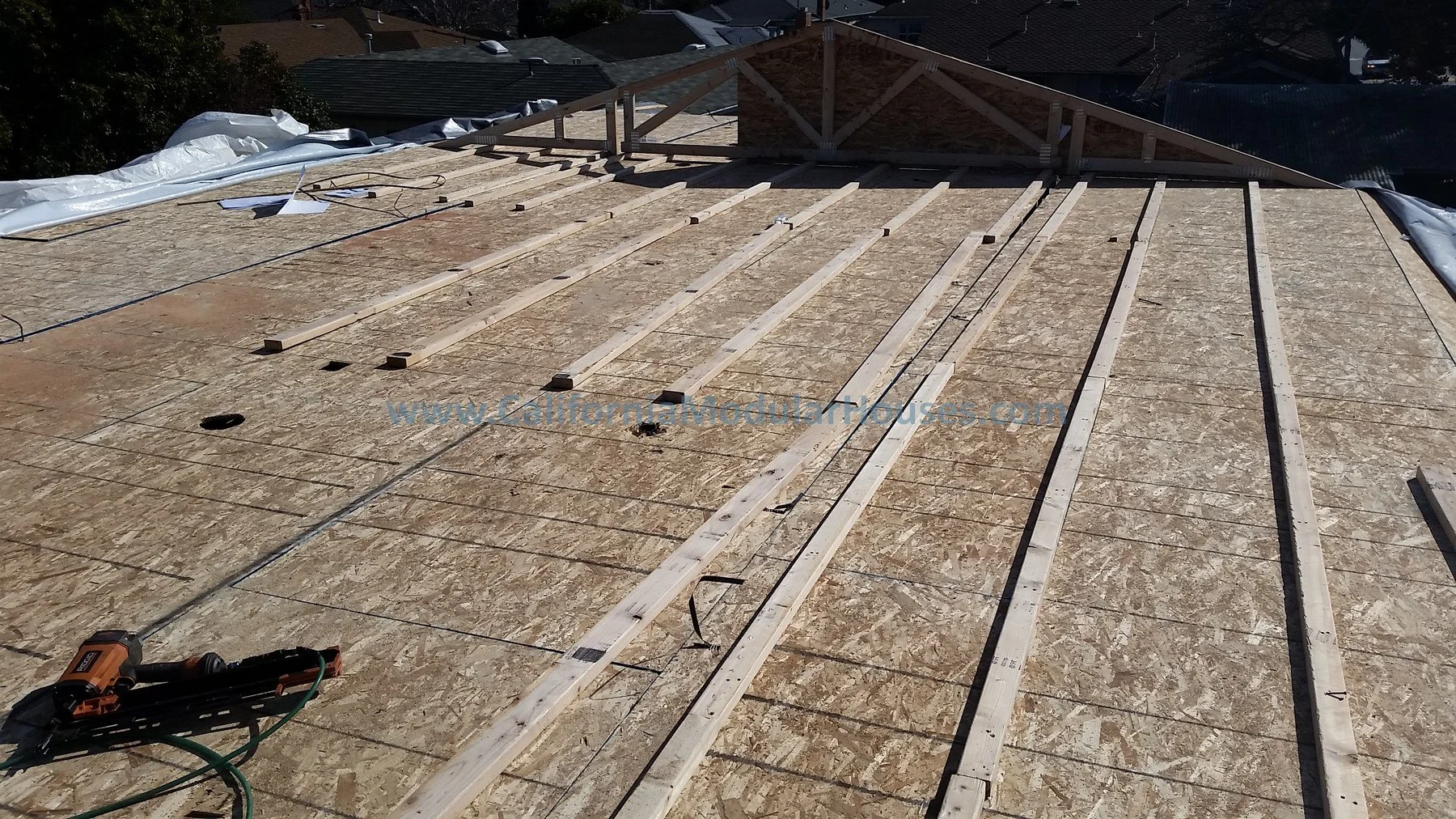 Construction site with wooden framing and OSB sheathing being installed on a roof, with tools and materials scattered around. San Mateo Modular Home, California Modular Home.
