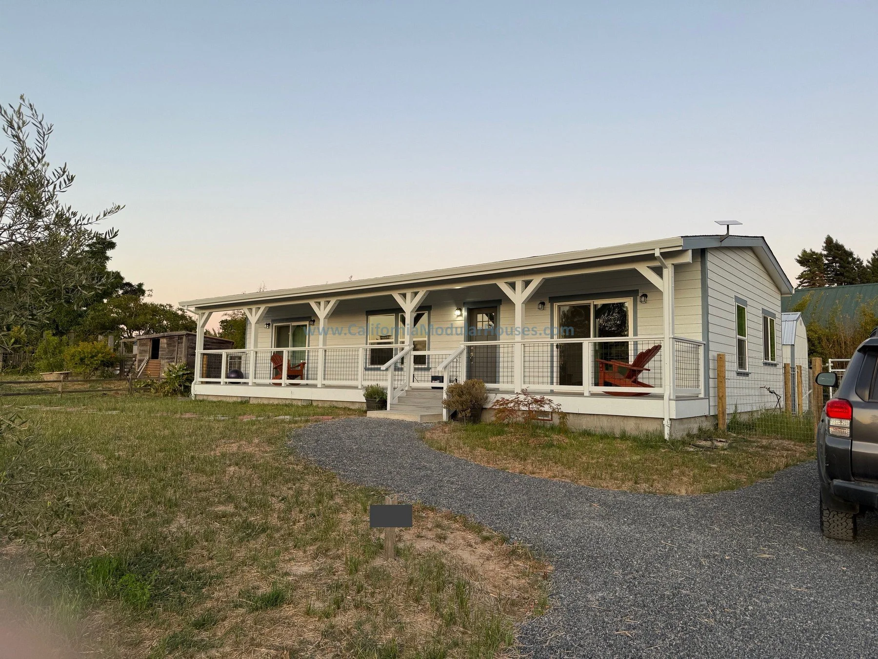 A modern, single-story white house with a porch, outdoor seating, and a gravel pathway leading to the front door, surrounded by grass and trees.  Sebastopol, Sonoma County, CA.  Prefab modular Accessory Dwelling Unit.  