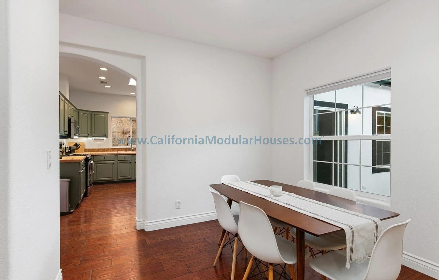 Modern dining area with a wooden table and six white chairs, adjacent to a large window, with a view of a patio. The space opens into a kitchen with green cabinets and wooden countertops, wood flooring, and a white ceiling.