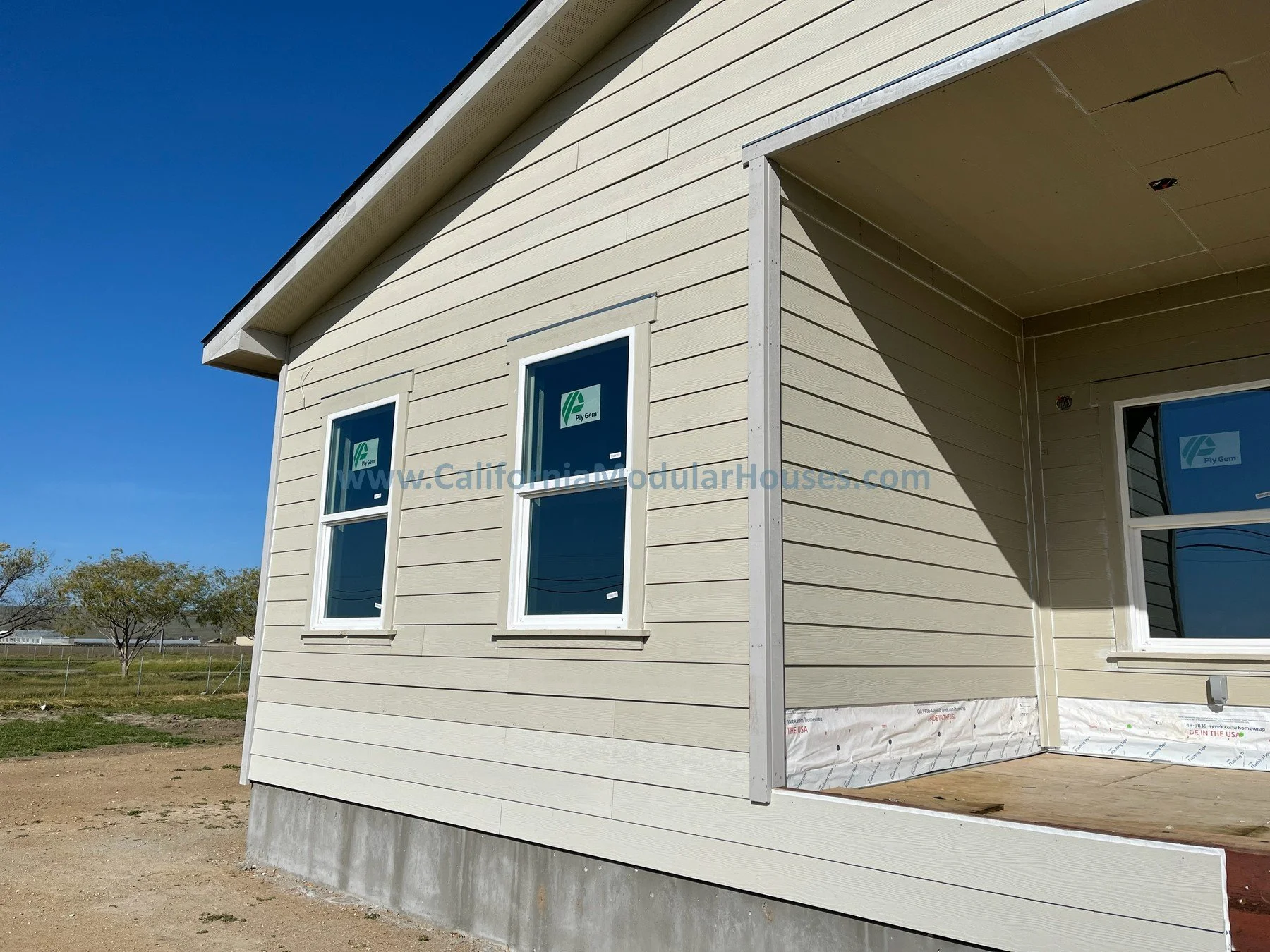 A beige, newly constructed modular house with three windows on the side. The house is built on a concrete foundation, and part of the porch is under construction. prefab home california.
