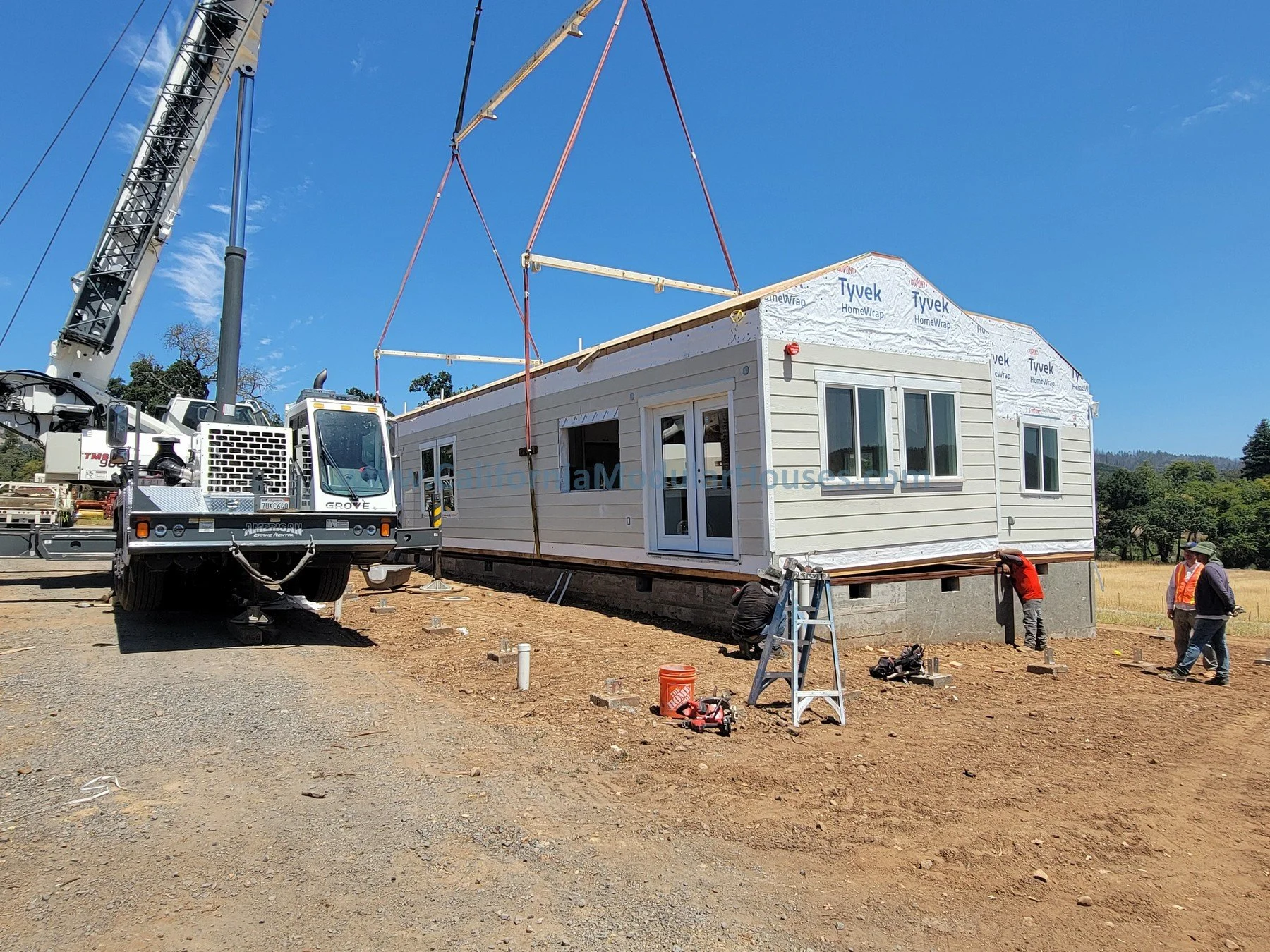 Construction workers and a crane installing a prefabricated house on a foundation at a construction site under a blue sky.