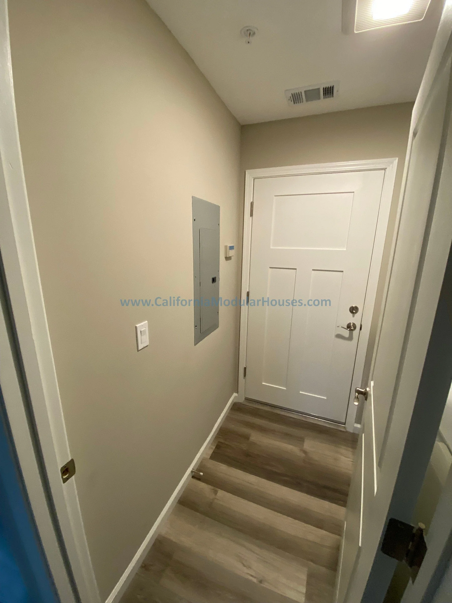 Neat entryway with a white door, electrical panel, thermostat, and wood-look flooring.   Modular Accessory Dwelling Unit  Modular ADU