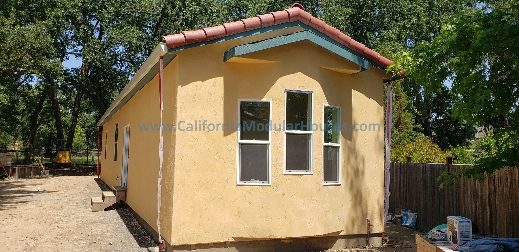 A newly constructed small yellow house with three windows on the front and a colorful sloped roof, situated in a yard with an ongoing construction area and green trees surrounding it.  Prefab Modular Accessory Dwelling Unit.  