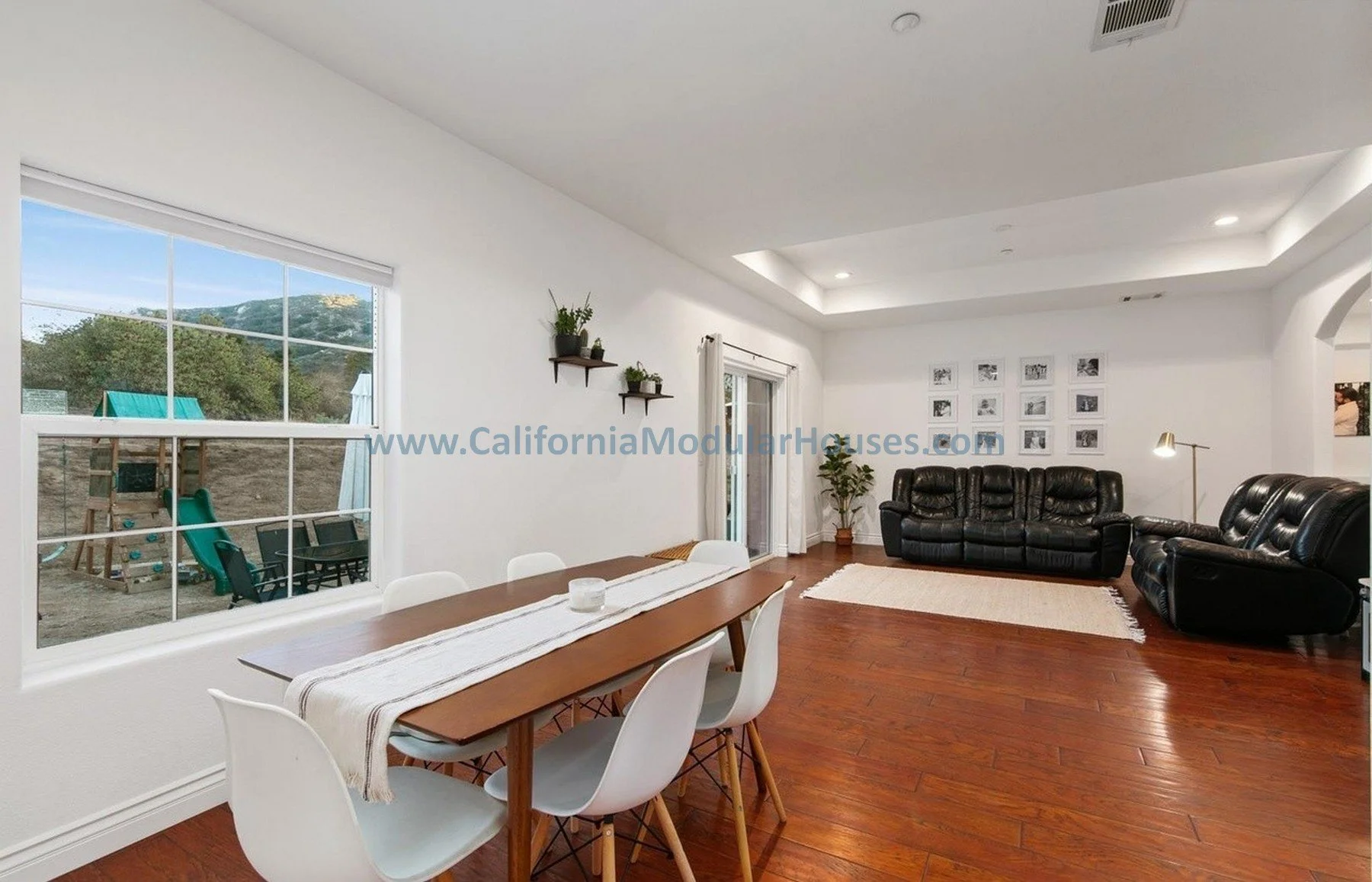 Living room with large window, black leather sofas, wooden floor, wall art, and a dining table with chairs.