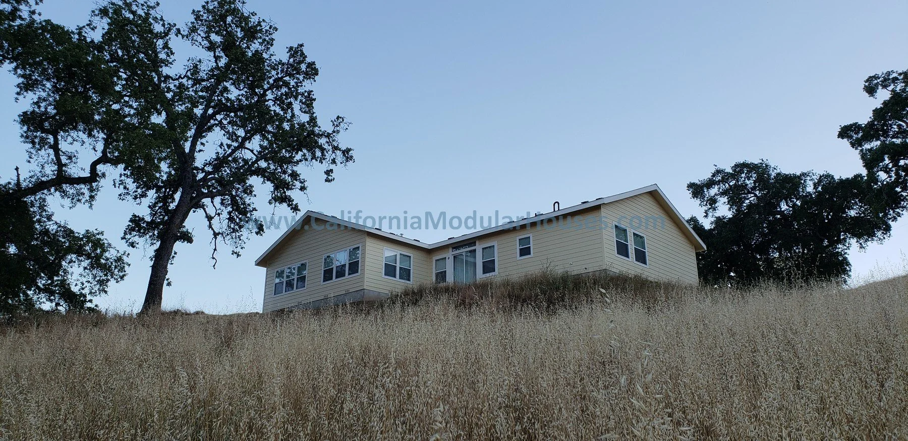A two-story beige house on a hill with dry grass and large trees, under a clear blue sky.  Bay Area Prefab Homes, California Modular.