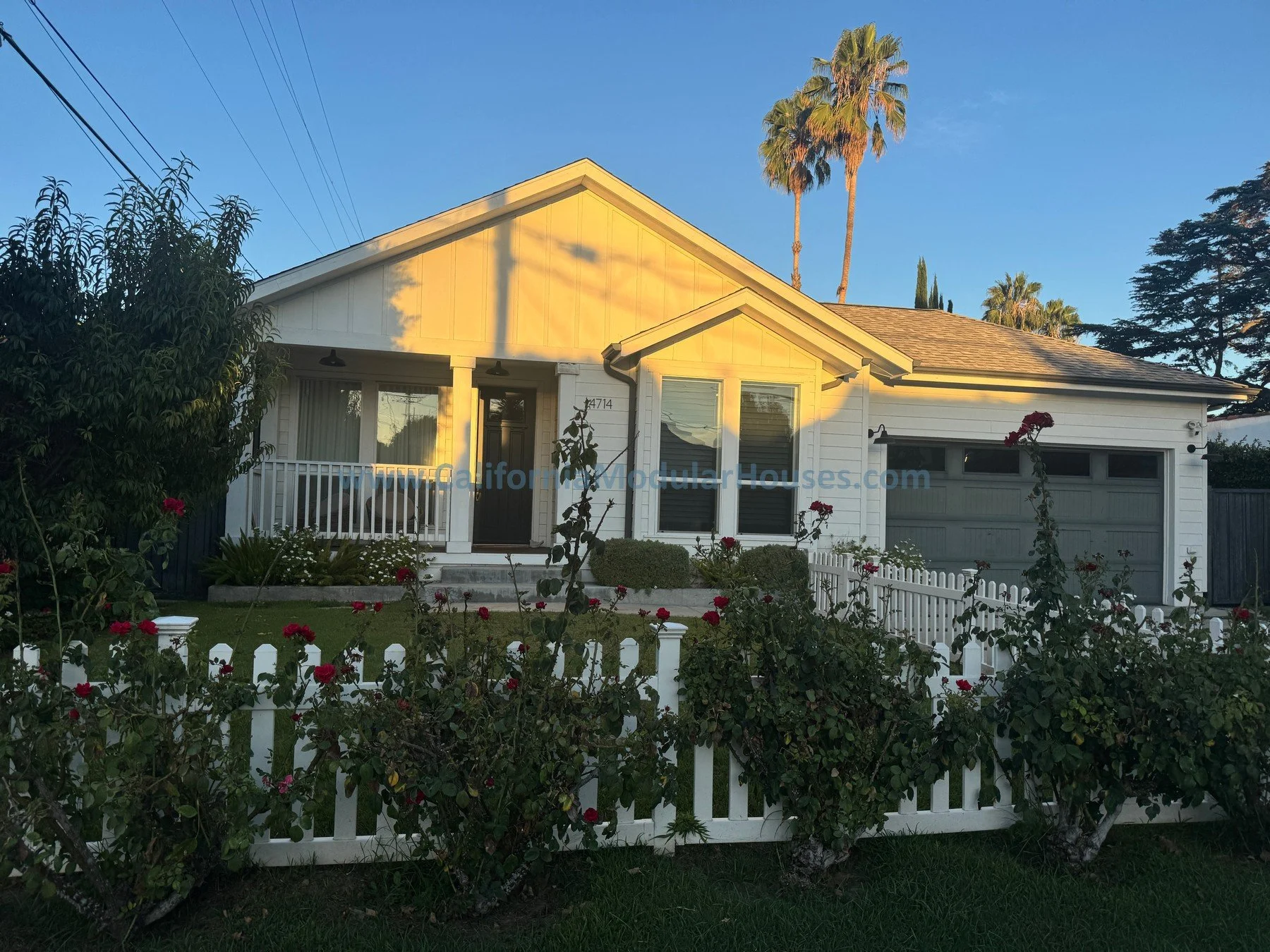 A white house with a front porch, surrounded by a white picket fence. There are blooming roses in the garden, tall palm trees in the background, and the sky is a clear blue with the warm glow of sunlight.