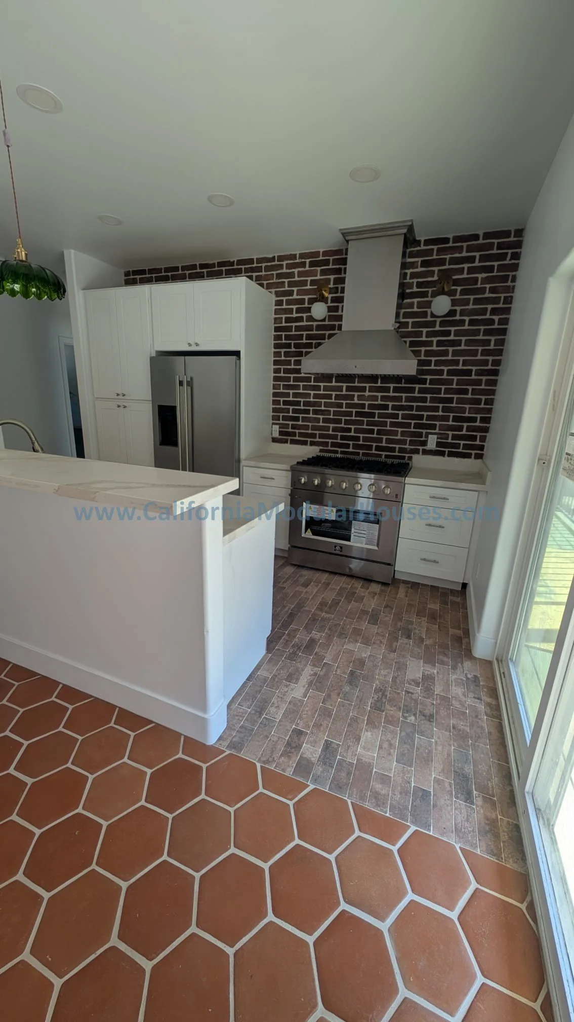 Kitchen with terracotta hexagon floor tiles, brick accent wall, stainless steel oven and refrigerator, white cabinets, and a glass sliding door.